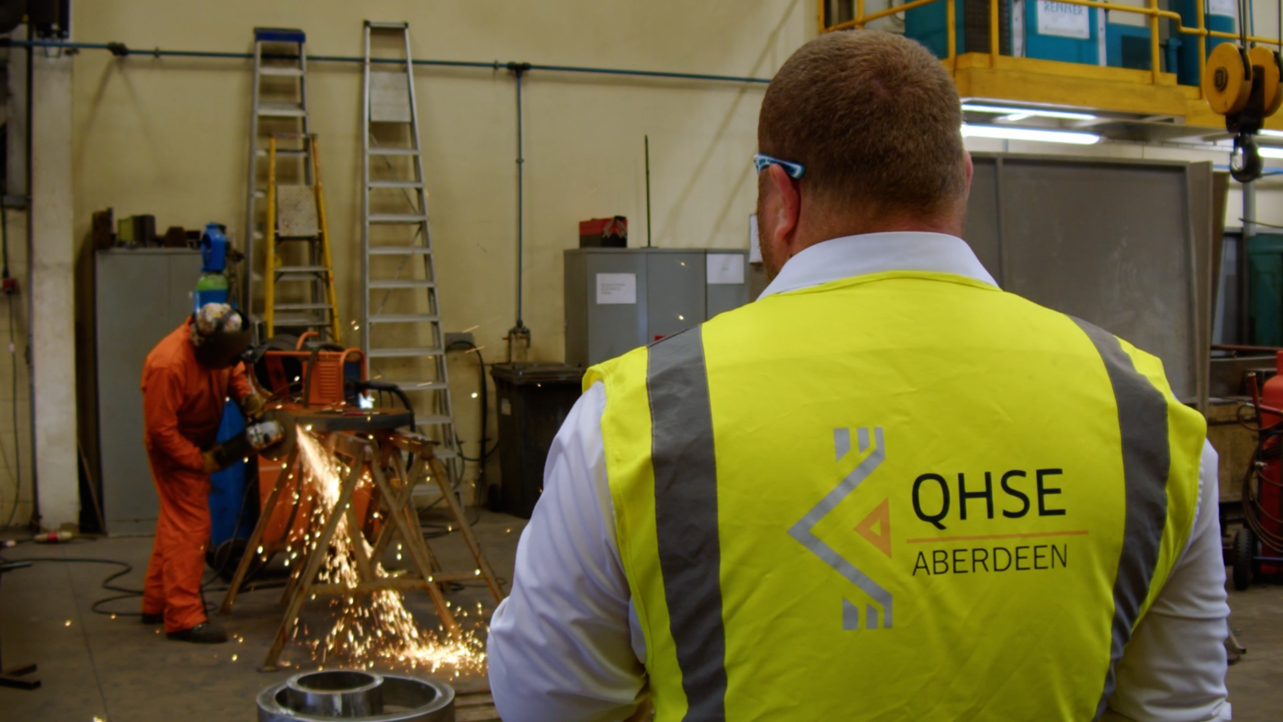 QHSE Aberdeen representative observing a worker using a saw tool inside an industrial warehouse.
