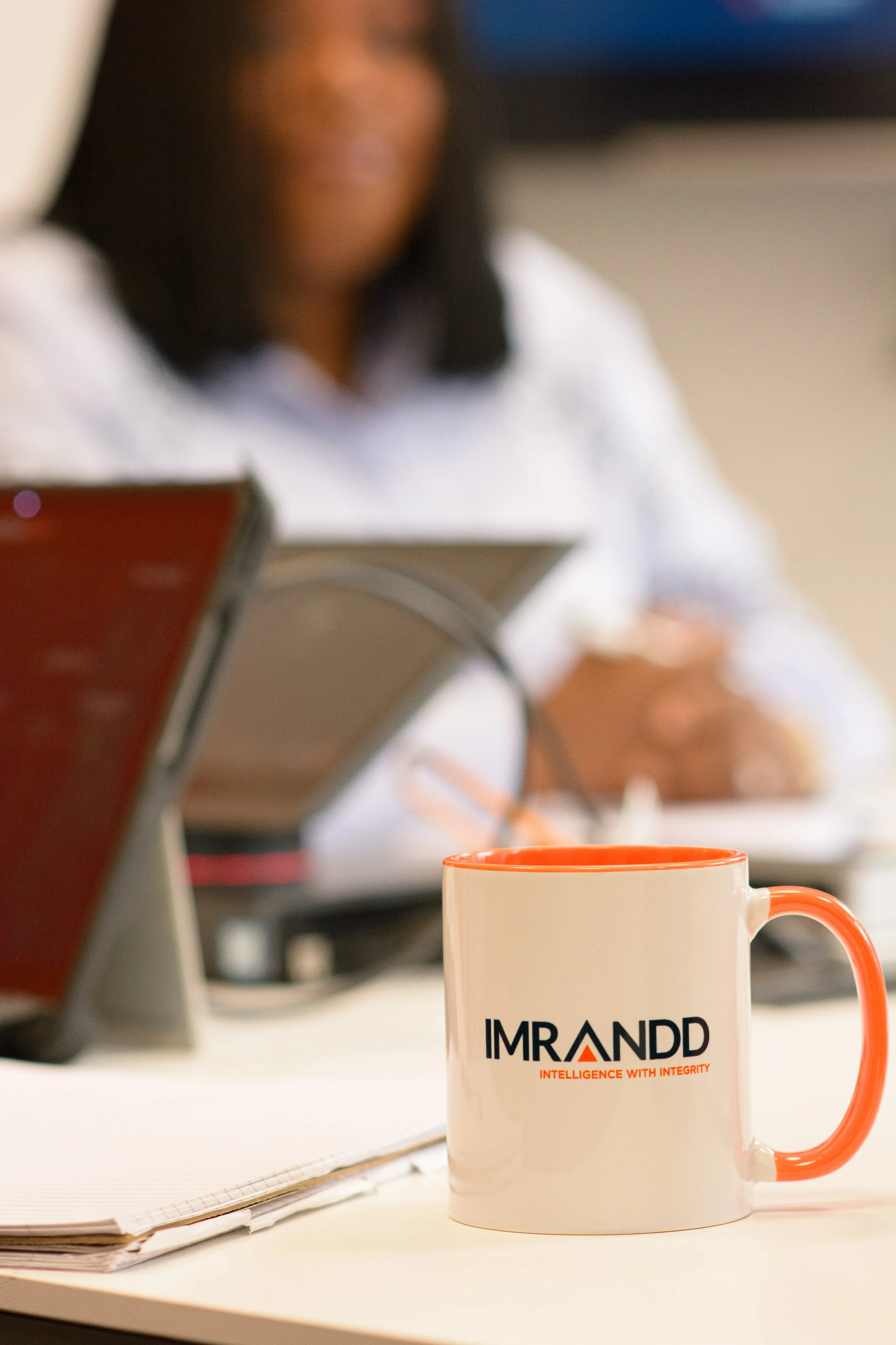IMRANDD branded coffee cup on a table with a team member blurred in the background.