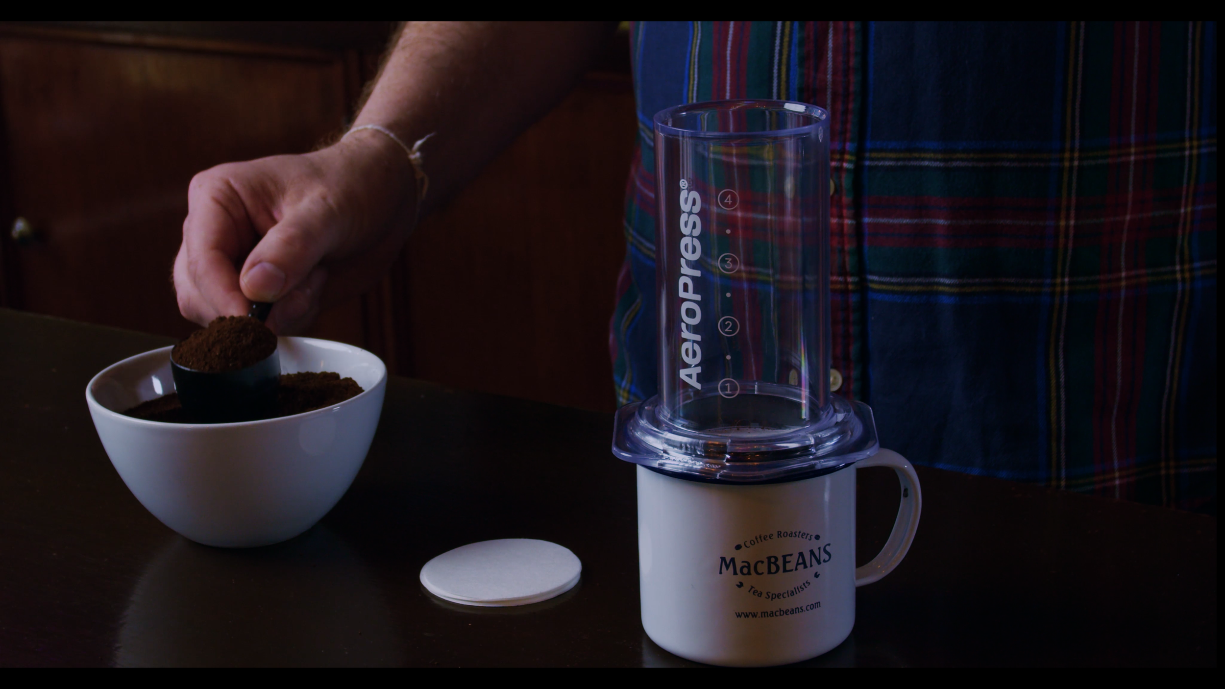 A man scooping ground coffee into an AeroPress above a MacBeans cup.