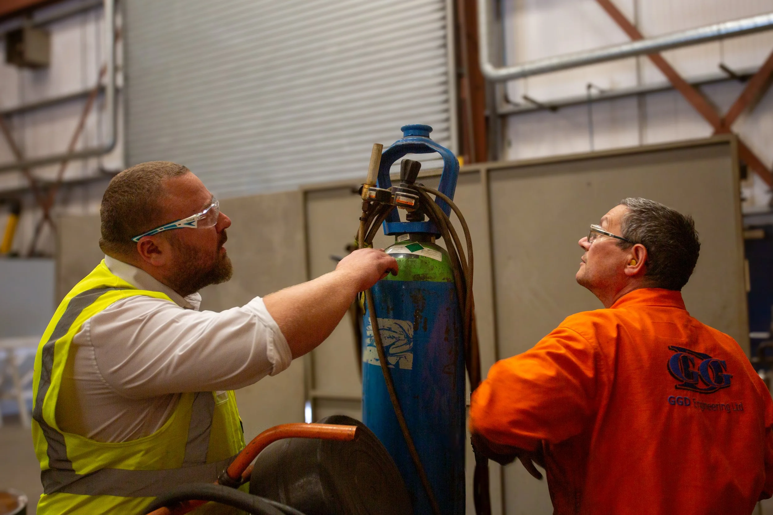 QHSE Aberdeen team member observing a worker inspecting a canister inside an industrial warehouse.