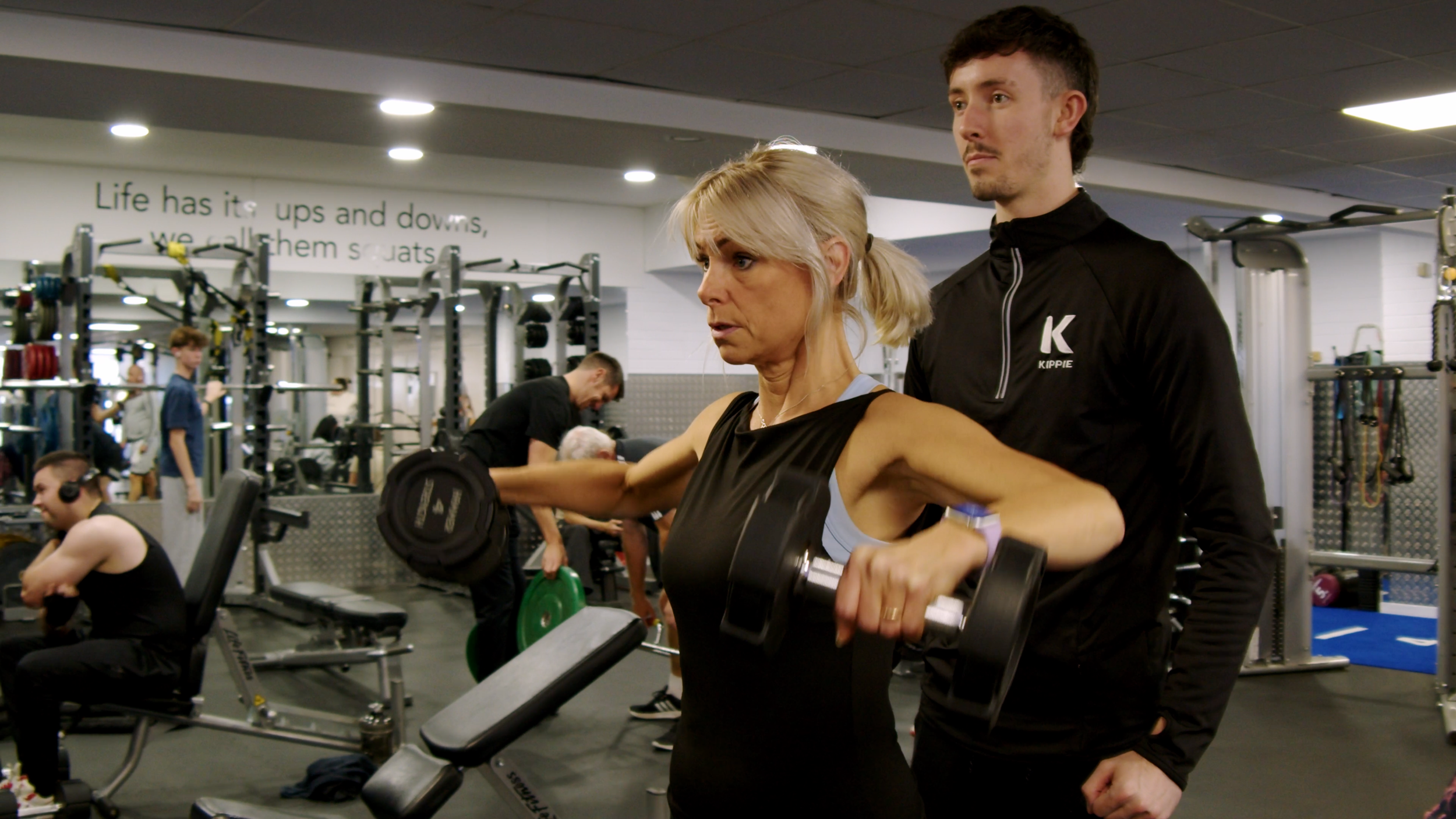 Woman lifting a weight while a coach watches inside the busy Kippie Lodge gym.