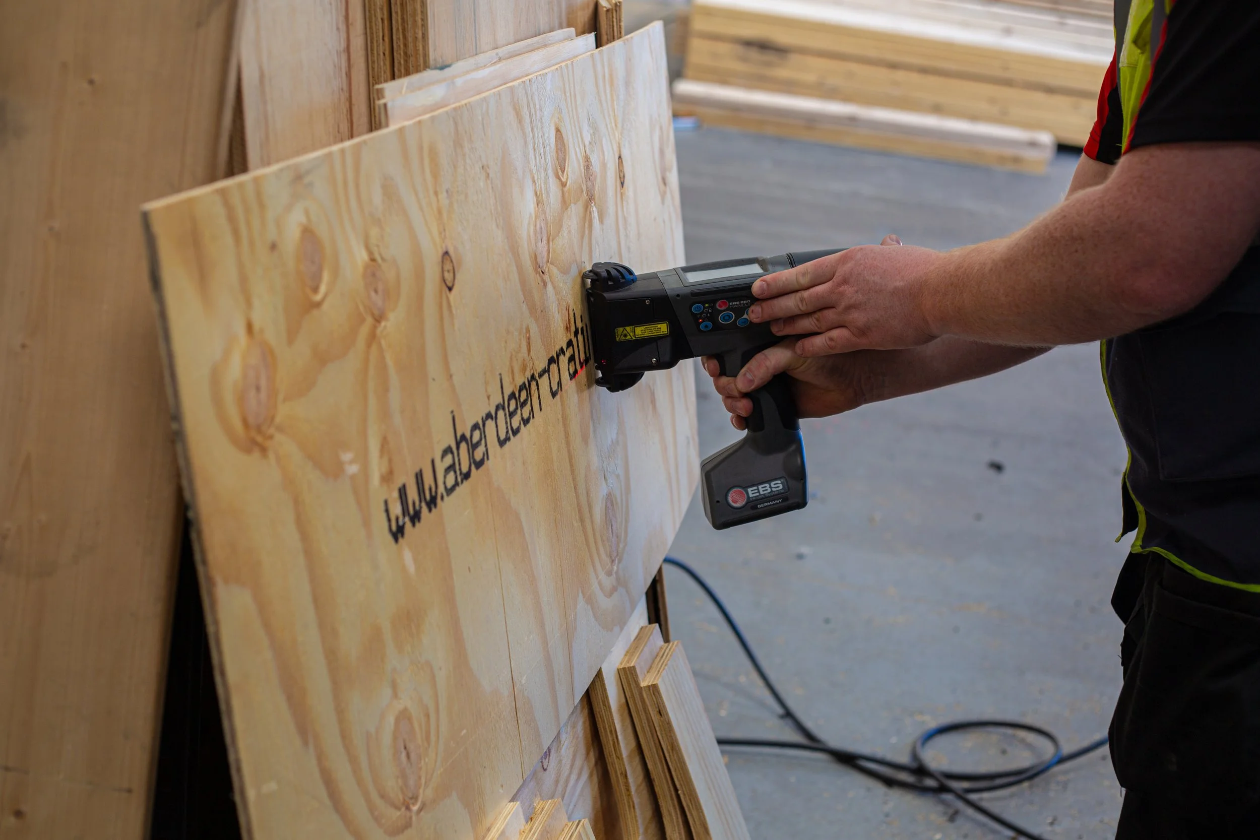 AIC worker using a handheld digital printer to mark the company website on plywood inside the warehouse.