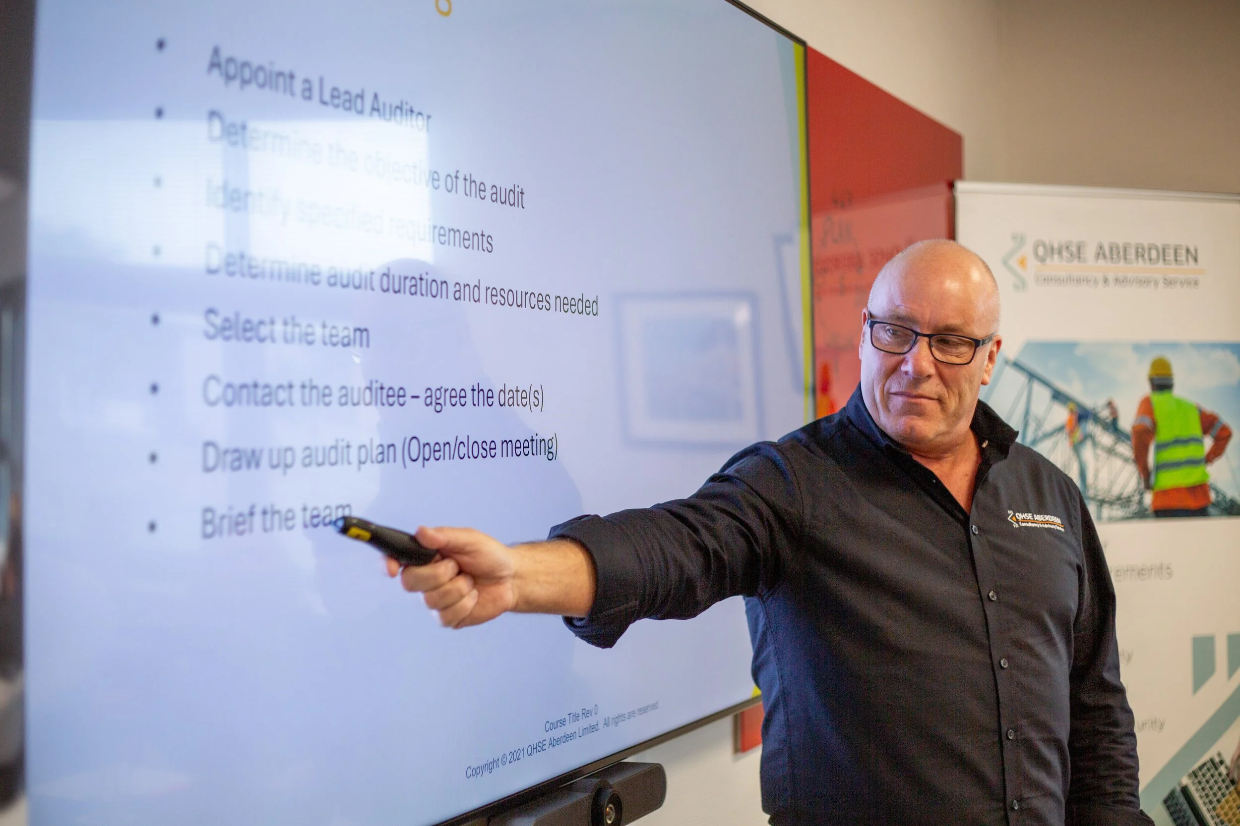 QHSE Aberdeen team member delivering a presentation and pointing to a briefing displayed on screen.