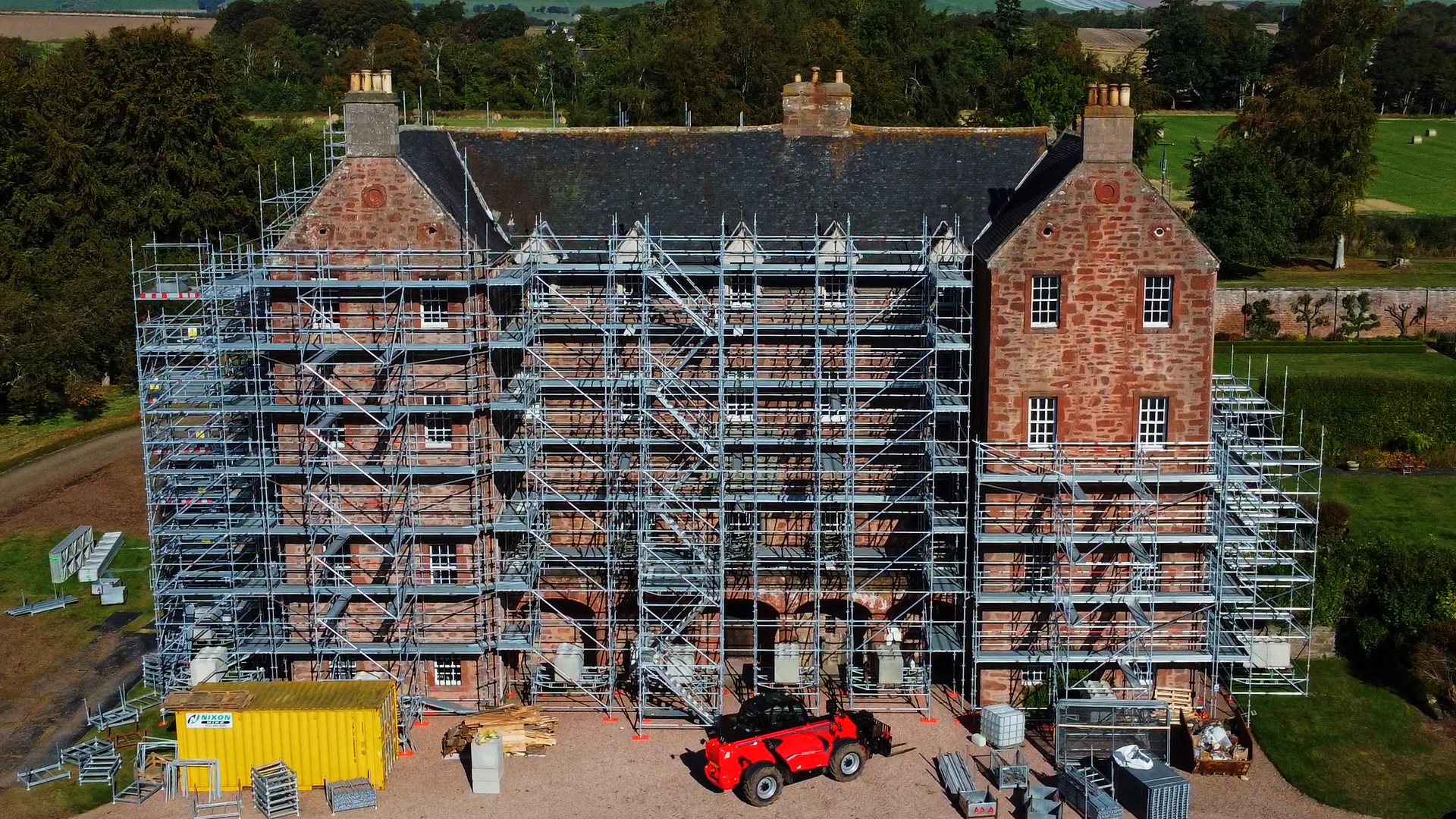 Large residential property surrounded by scaffolding with a forklift, yellow container and equipment around the site.