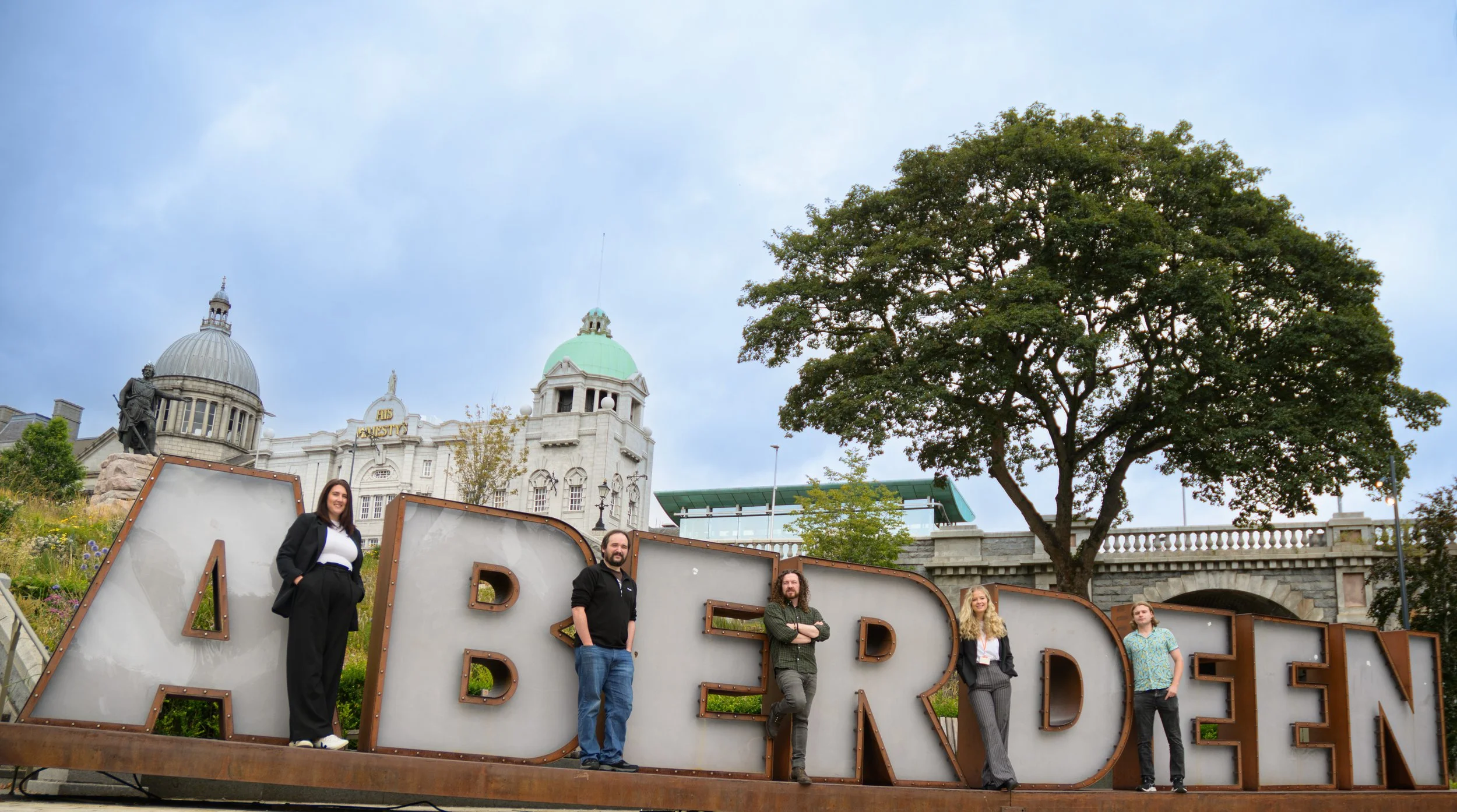 IMRANDD team standing beside the Aberdeen sign in Union Terrace Gardens with His Majesty’s Theatre behind.