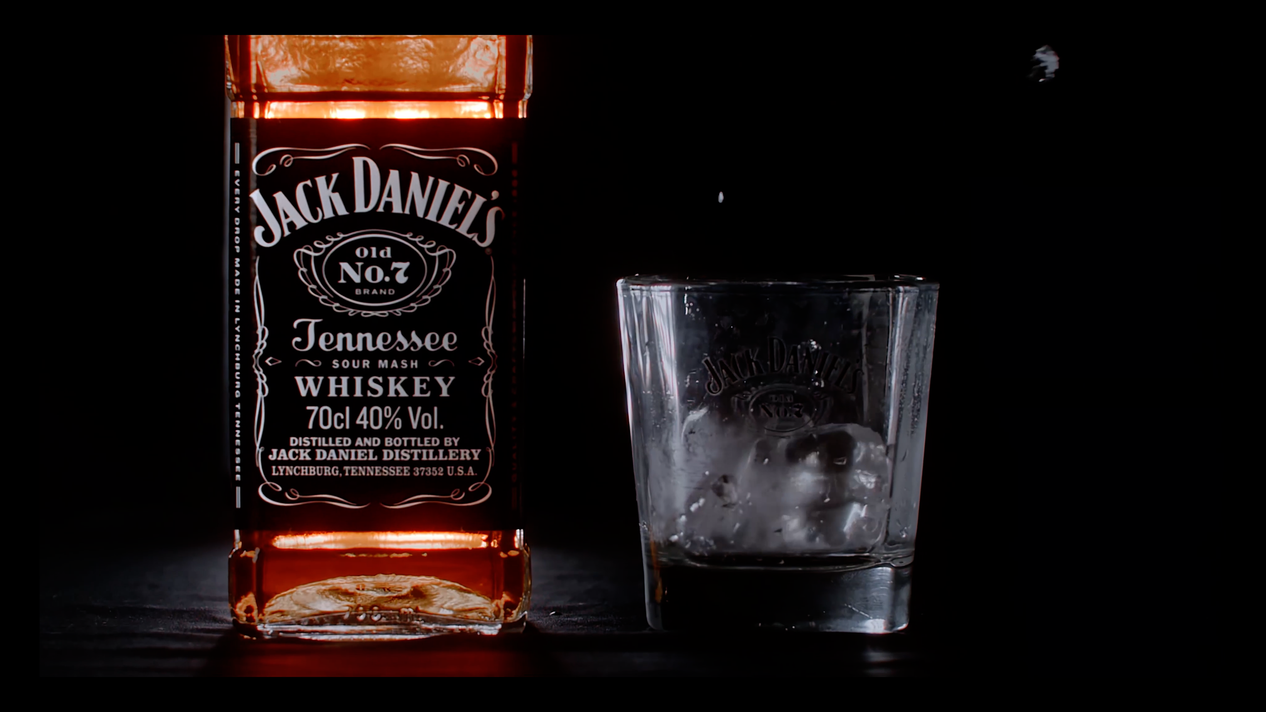 Close-up of a Jack Daniel’s bottle beside a glass as ice drops into it.