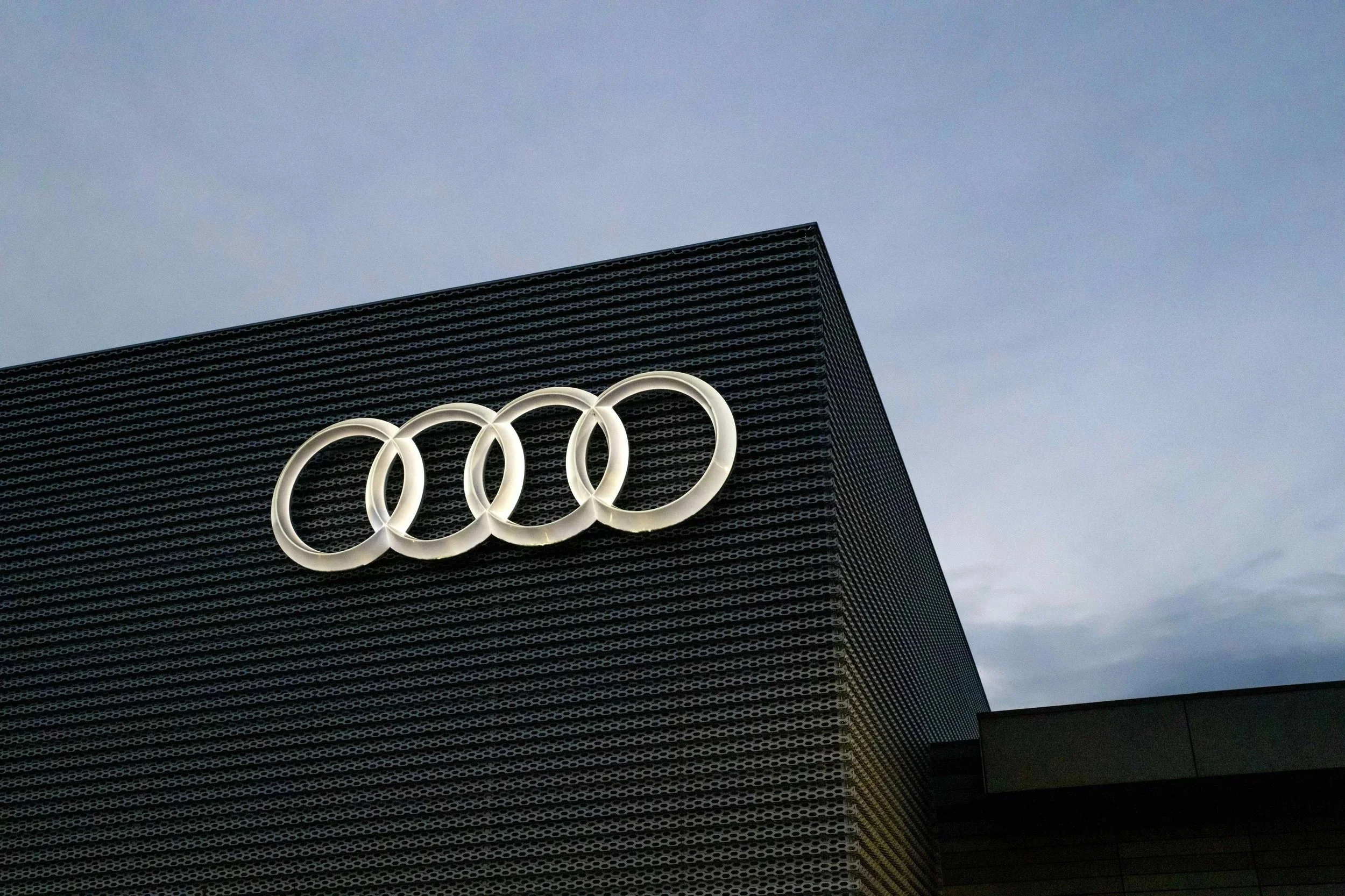 Exterior of a building with the Audi logo, a cluster of four interlinked silver rings, attached on a grid-like facade under an overcast sky.