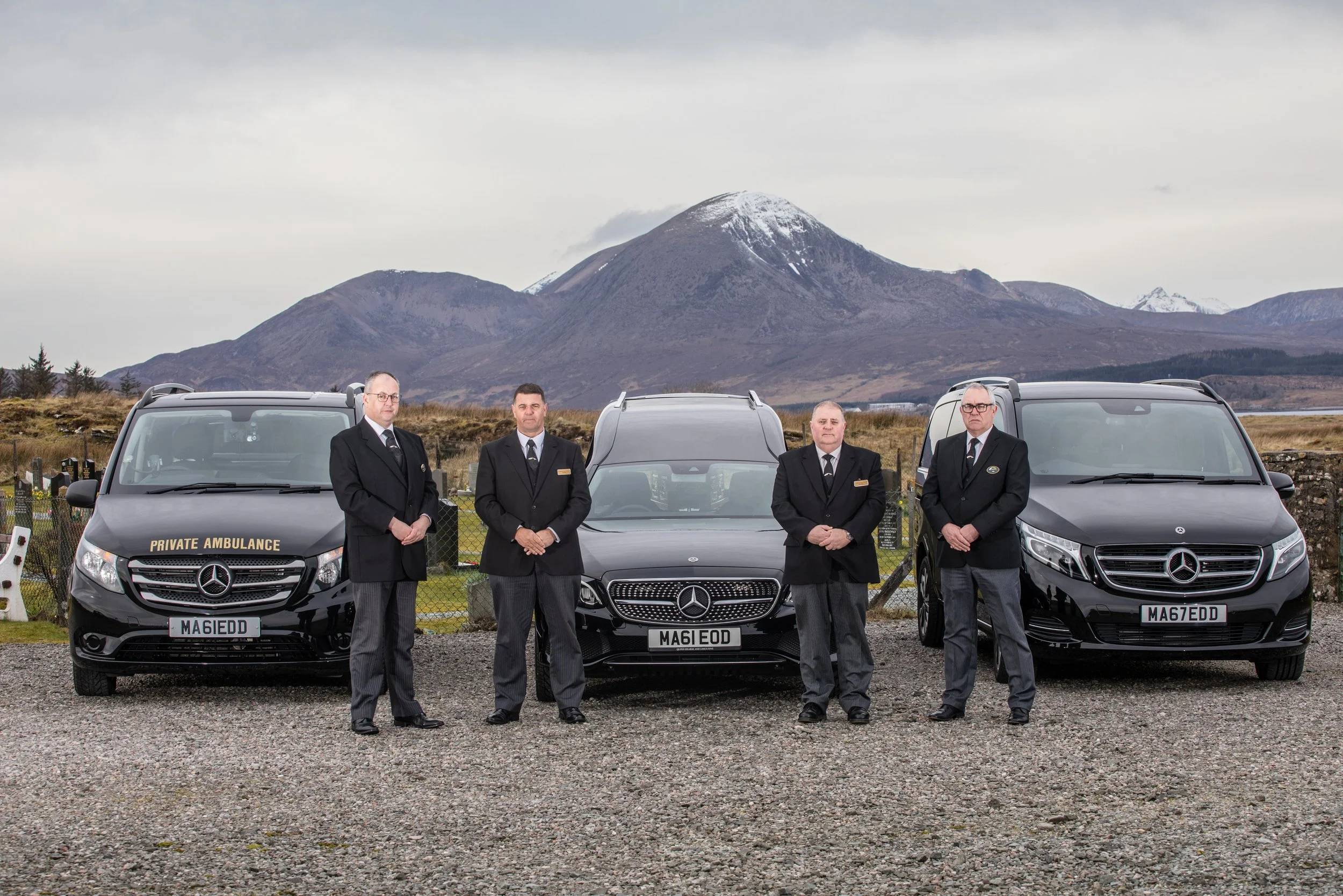 Four men in black suits and ties standing in front of three Mercedes-Benz vehicles labeled 'Private Ambulance' with a mountain and cloudy sky in the background.