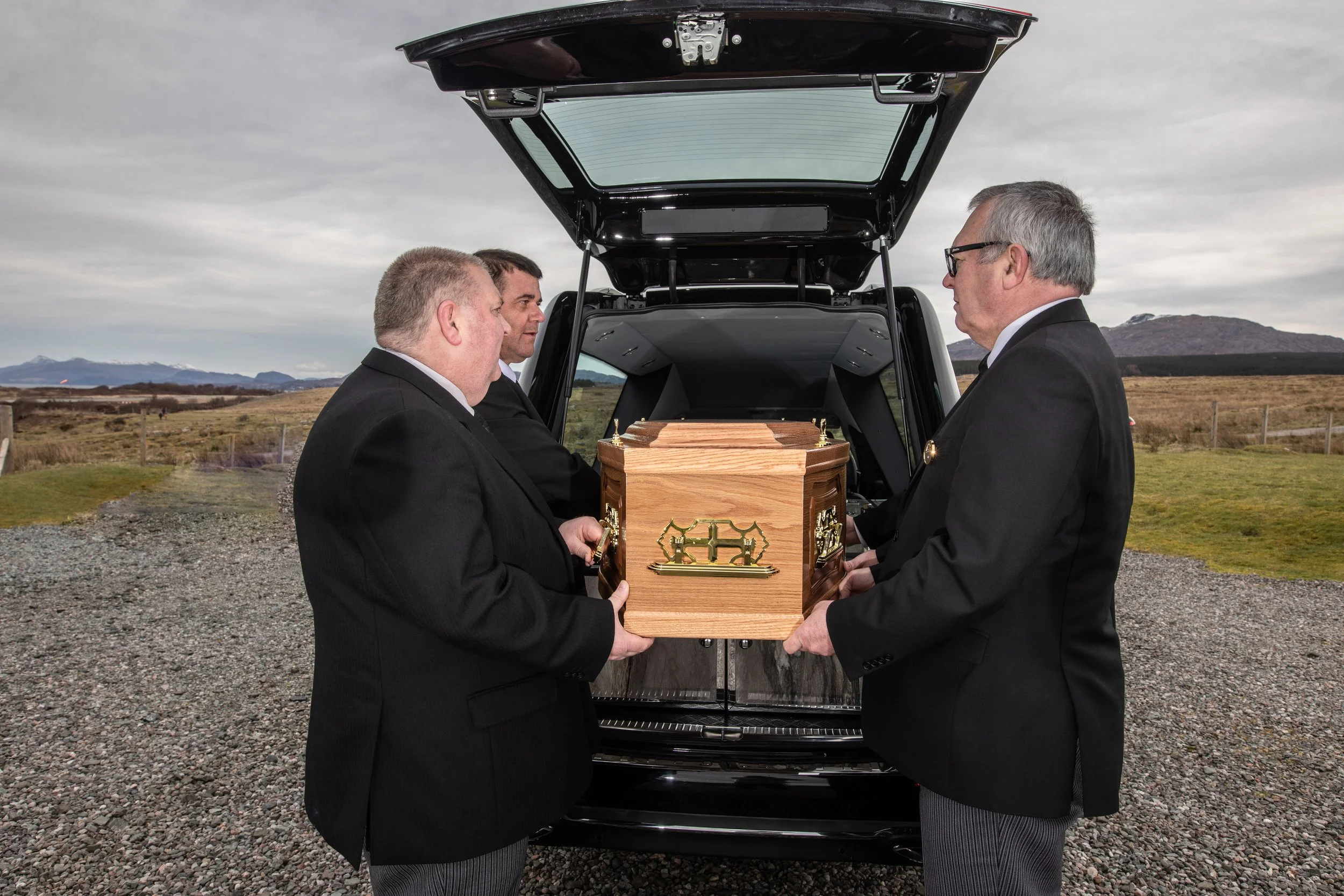 Three men in black suits and glasses holding a wooden coffin with gold accents outdoors near an open vehicle trunk, mountain scenery in the background.