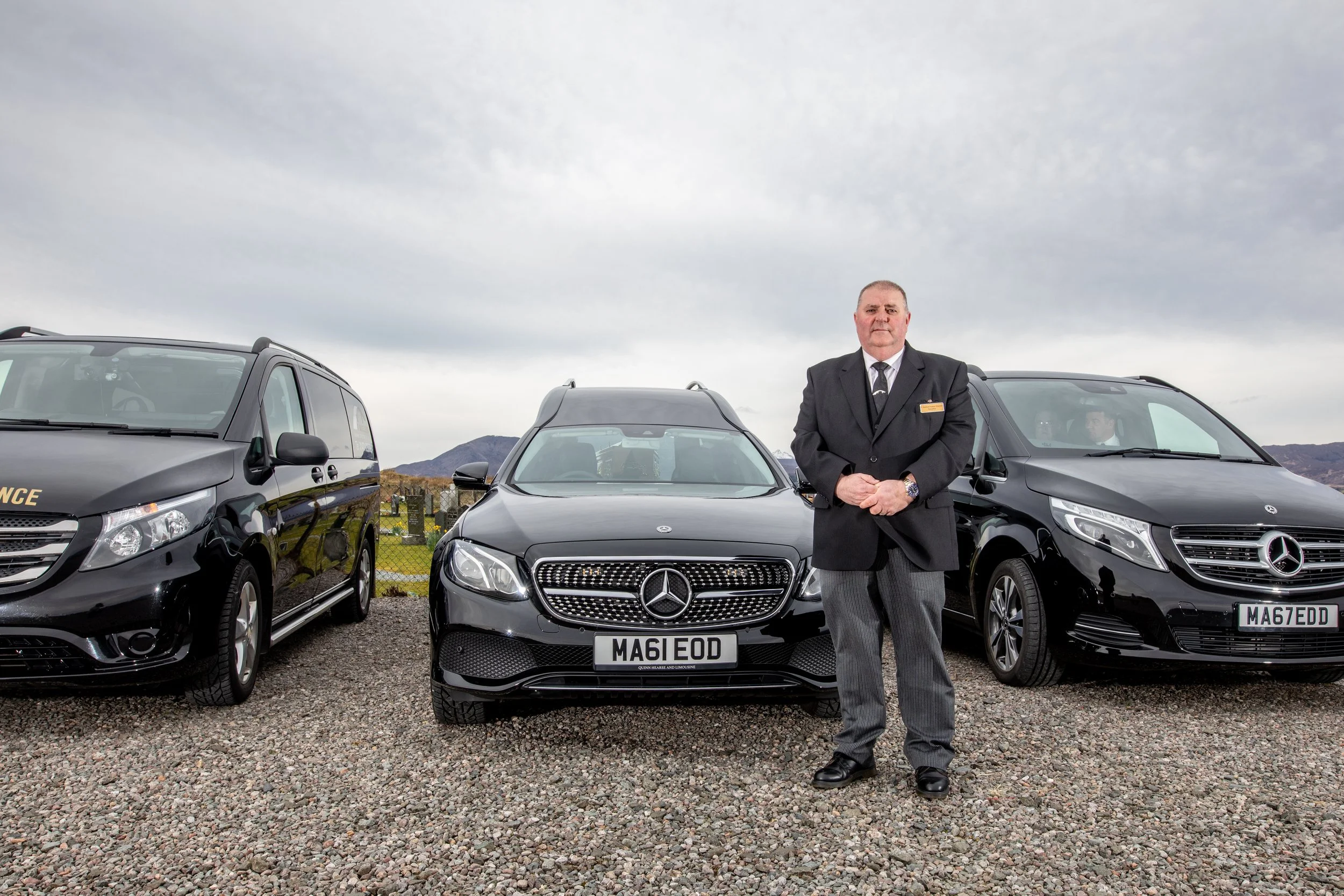 A man in a suit standing in front of three black luxury cars, parked outdoors on a gravel lot, with mountains and an overcast sky in the background.