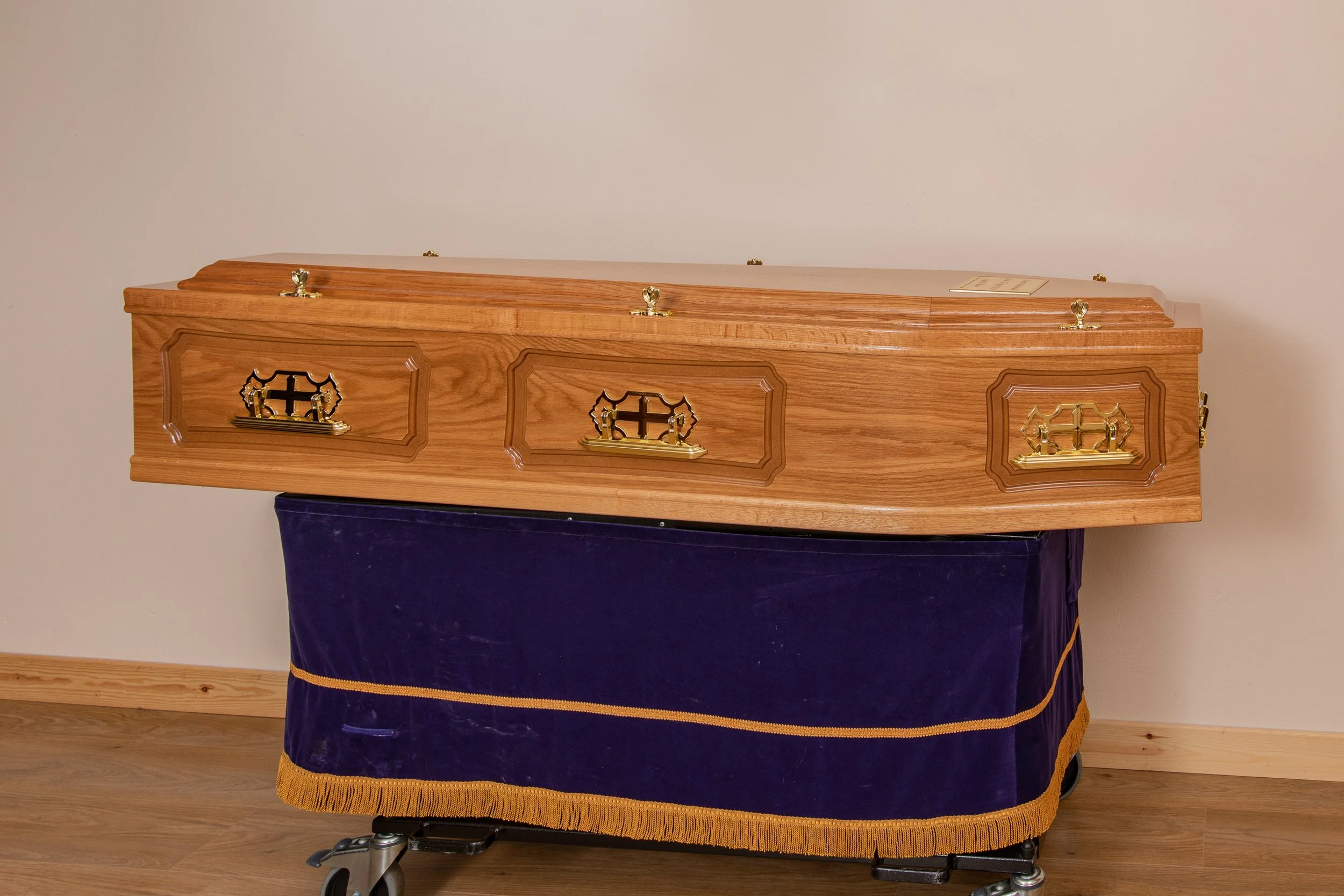 Raised lid panel side coffin light oak veneer. Also available in dark oak veneer. Full details: raised lid and panel sides, metal handles, engraved name plate, 8 cords and comes complete with engraved grave marker as well as purple coffin cover to pr