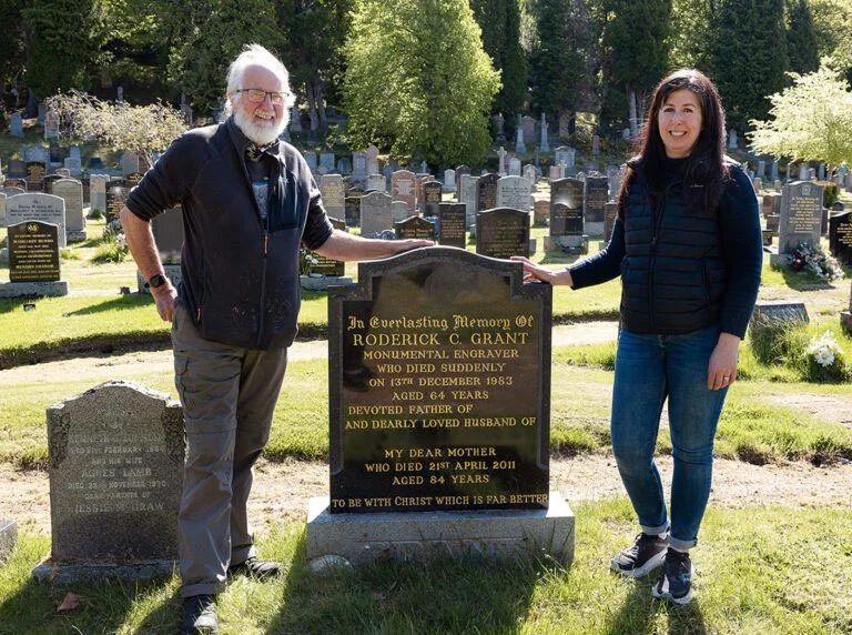 A man and woman standing on each side of an engraved grave marker in a cemetery, smiling. The grave marker commemorates Roderick C. Grant, who died in 1983 at age 64, and mentions his devotion as a father and husband, with an additional message about being with Christ.