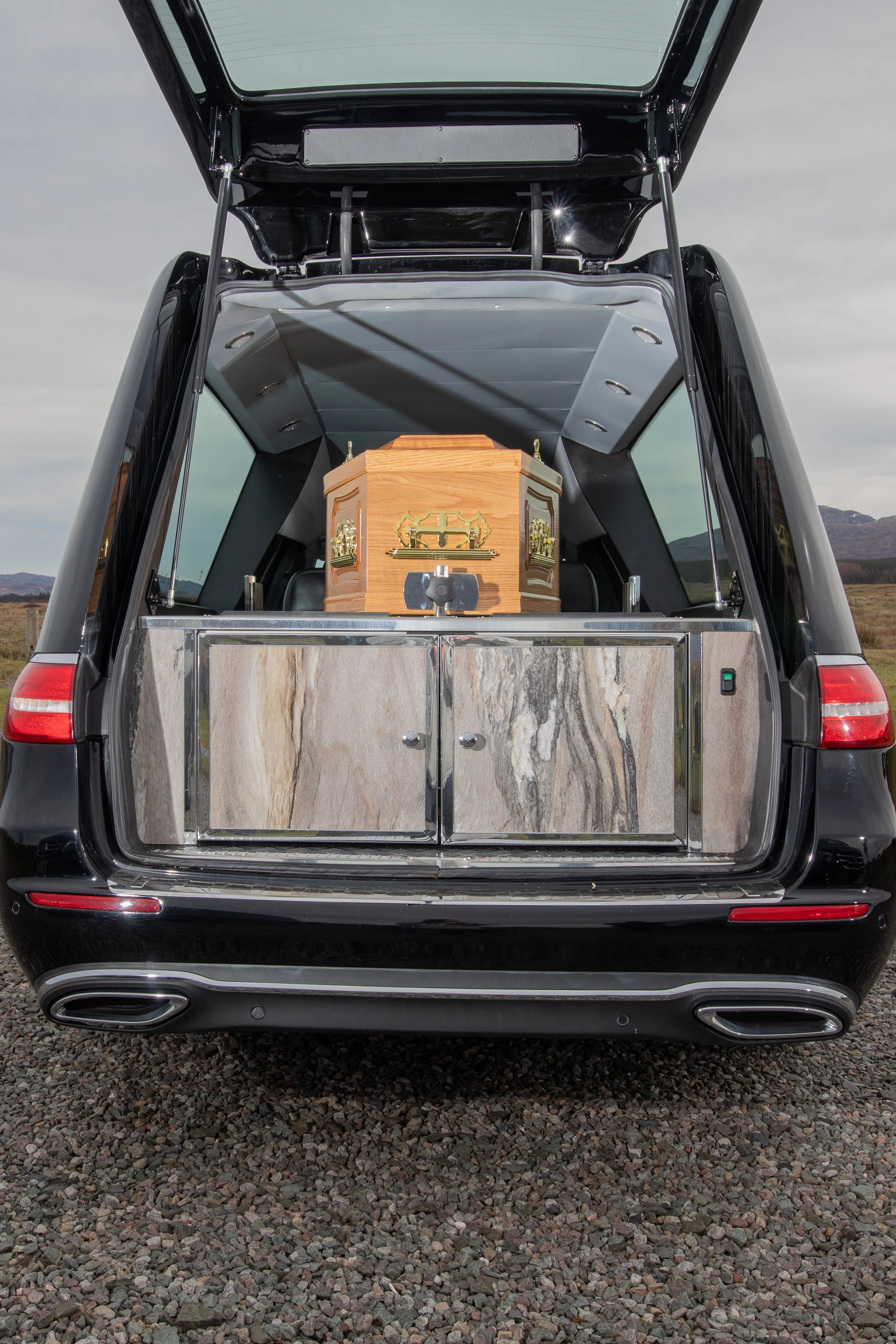 View of an open car trunk with a wooden coffin inside, parked on gravel with a landscape and mountains in the background.