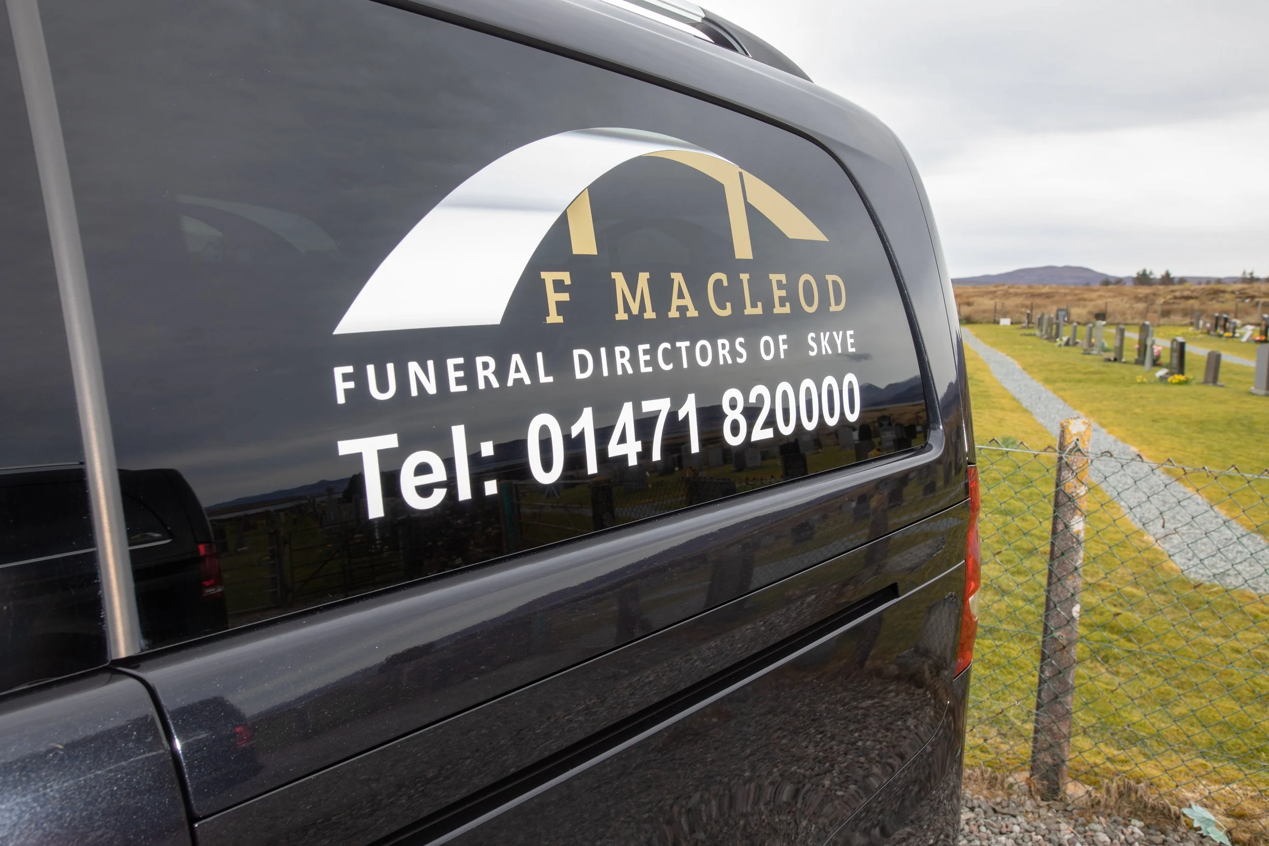 A black funeral vehicle with a sign reading 'F. Macleod Funeral Directors of Skye' and a phone number, parked near a cemetery with headstones, fencing, and a rural landscape in the background.
