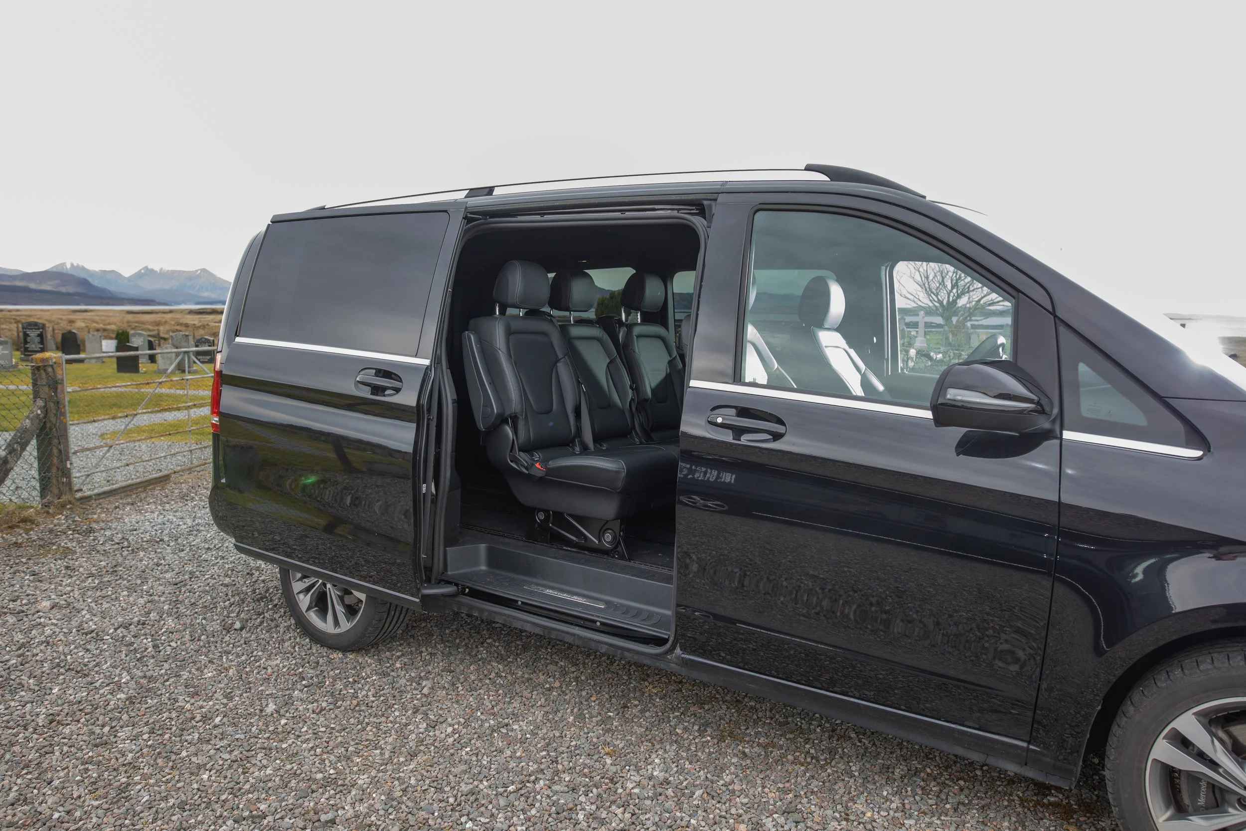 A black van with sliding door open, showing black leather seats inside, parked on gravel with a rural landscape and mountains in the background.