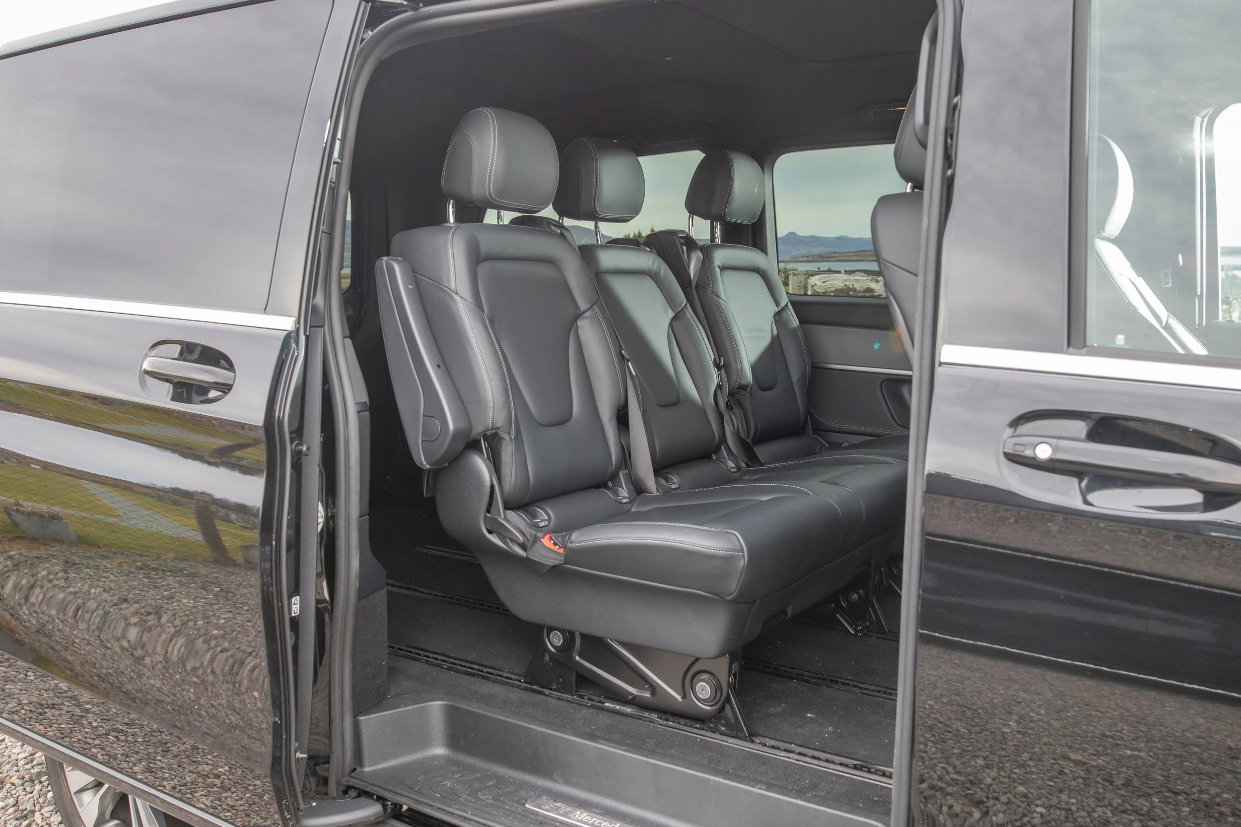 Inside a black van showing three rows of black leather seats with headrests, window scenery of mountains and a cloudy sky outside.