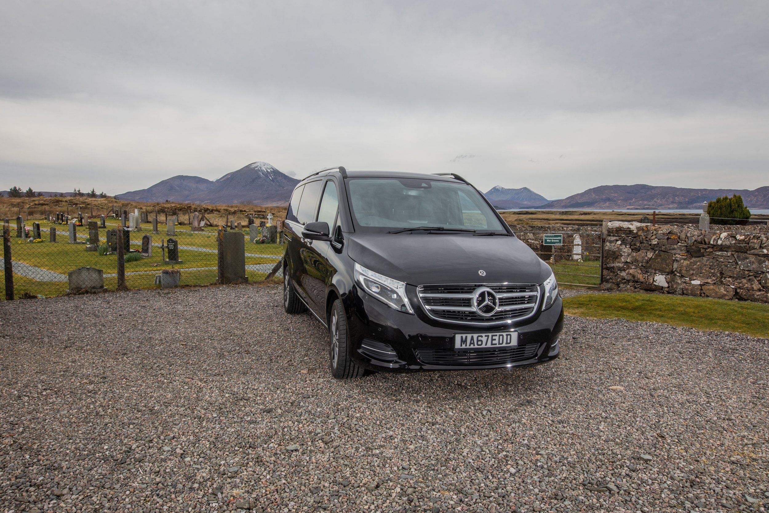 Black Mercedes-Benz van parked on a gravel surface near a graveyard with a mountain range in the background.