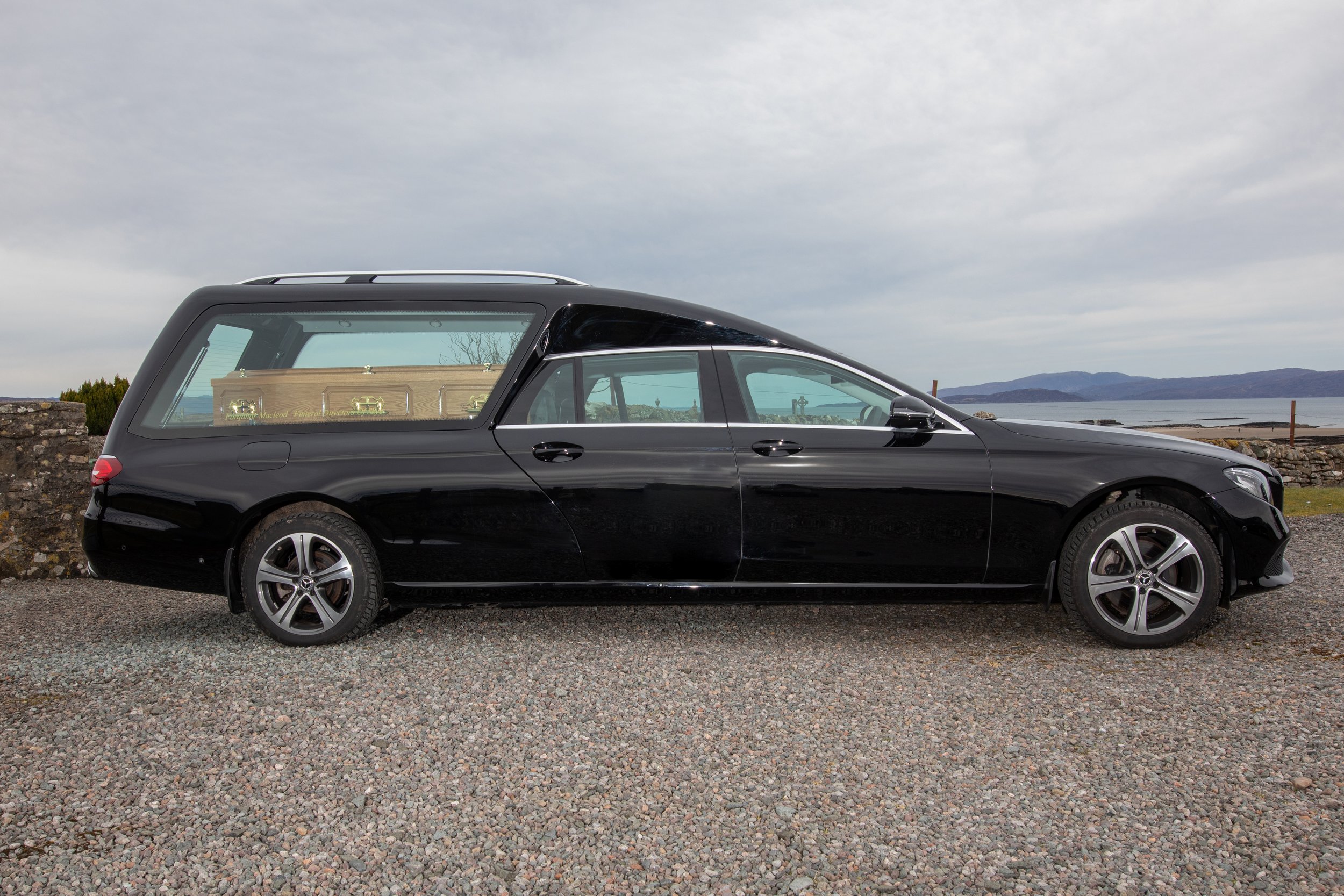 A black hearse parked on a gravel lot near a stone wall with a body in a casket visible through the glass window. Lake and mountain scenery in the background.