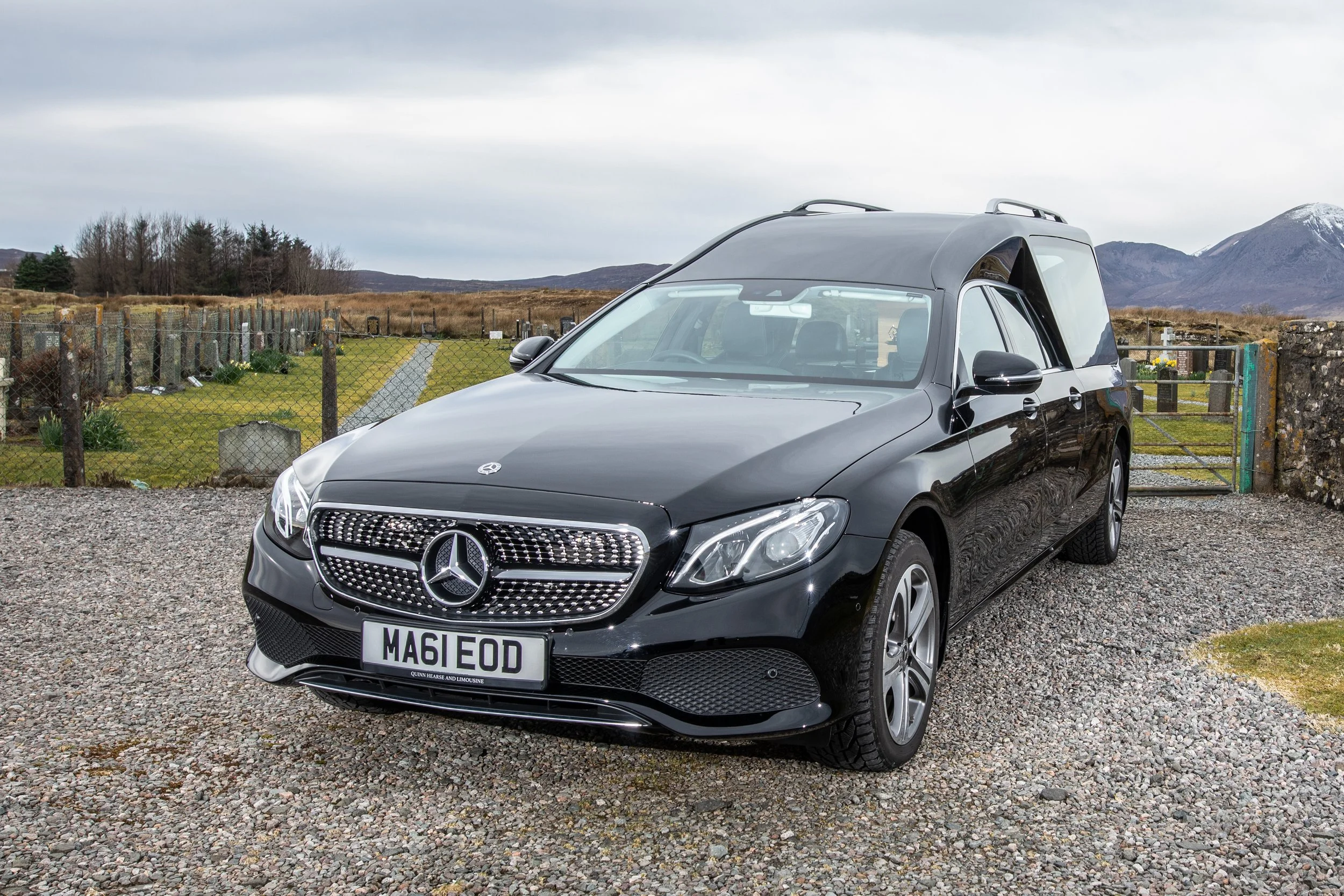 A black Mercedes-Benz car with a front grille displaying the Mercedes logo and a custom license plate that reads 'MA61 EOD', parked on a gravel surface near a cemetery with tombstones and a gate, against a backdrop of mountains and cloudy sky.