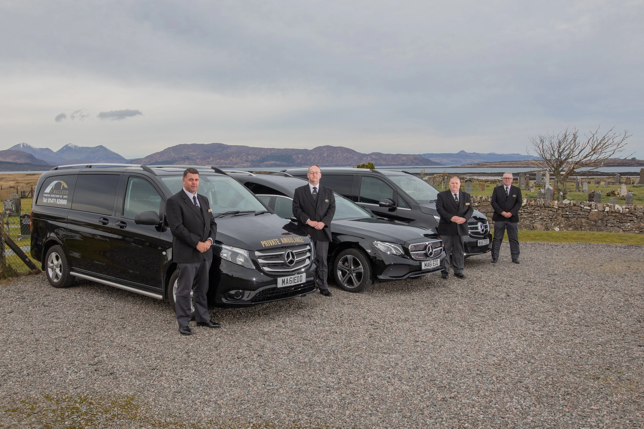 Four men in black suits standing in front of black Mercedes Benz vehicles, in an outdoor cemetery setting with mountains in the background. The vehicles are marked for a private ambulance service, and one vehicle displays a sign for "MacLeod Funeral 