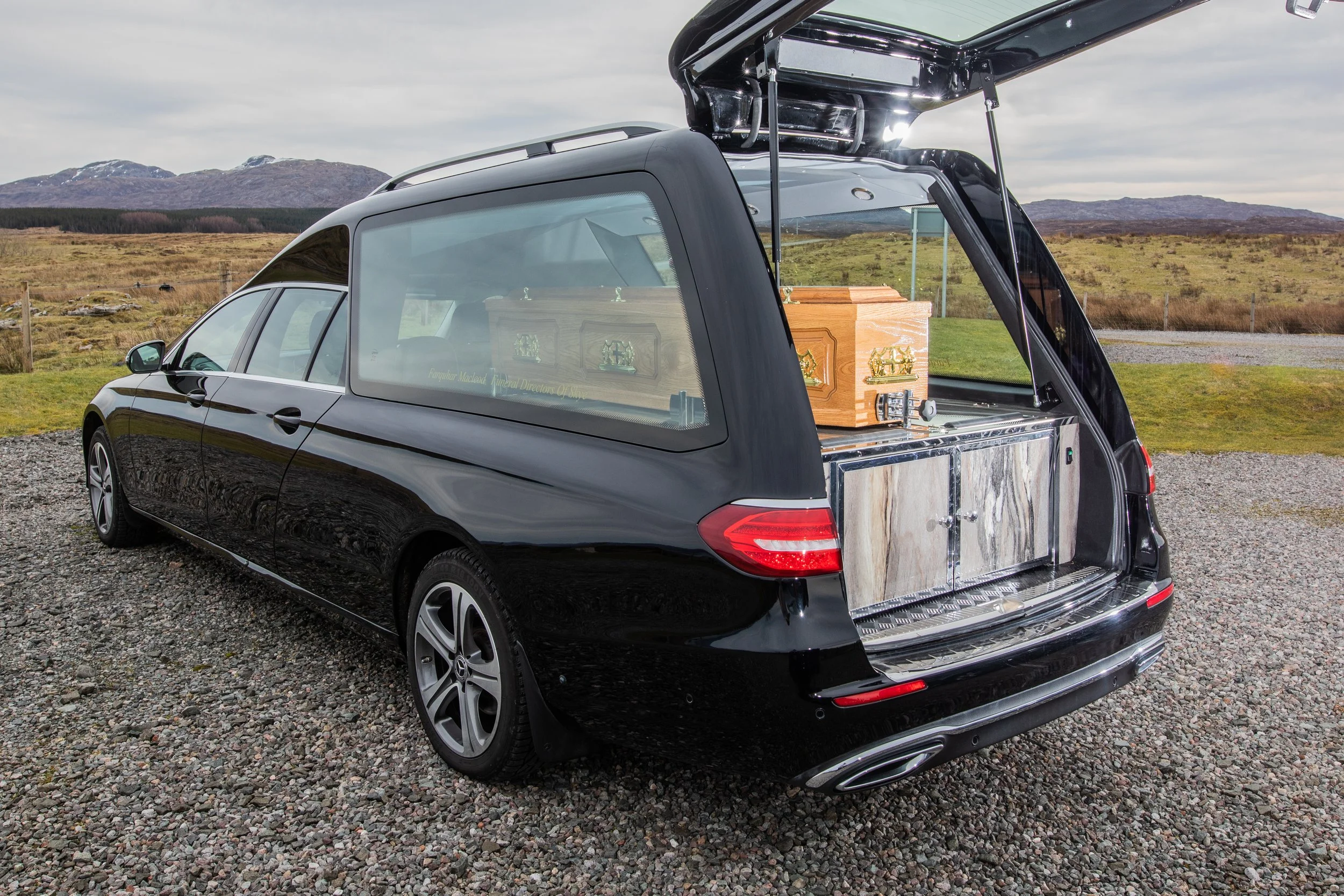 A black Mercedes-Benz car with its rear hatch open, showing a wooden coffin inside, outdoors in a rural area with mountains and fields in the background.