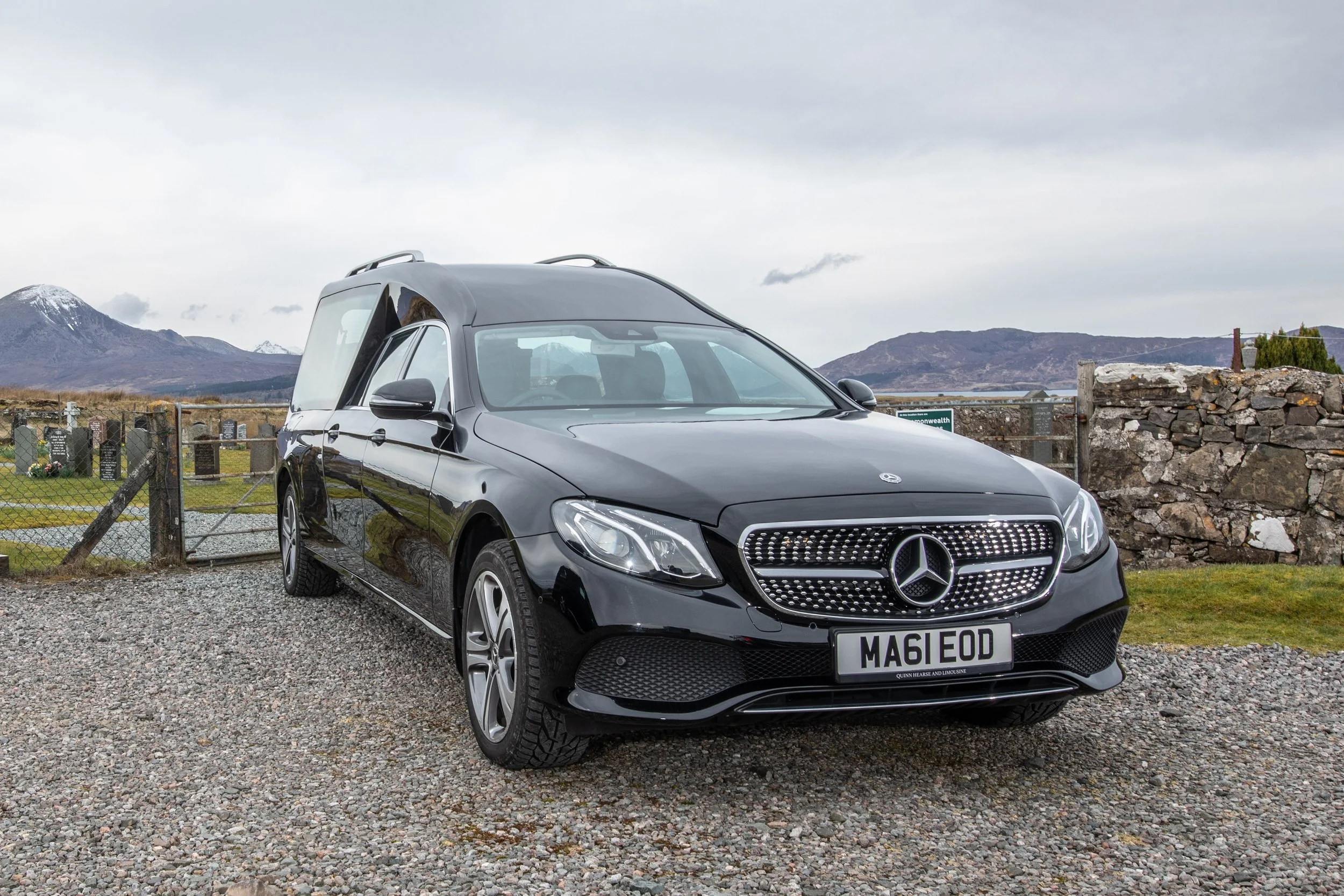 A black Mercedes-Benz car parked on a gravel surface in front of a stone wall, with mountains and cloudy sky in the background.