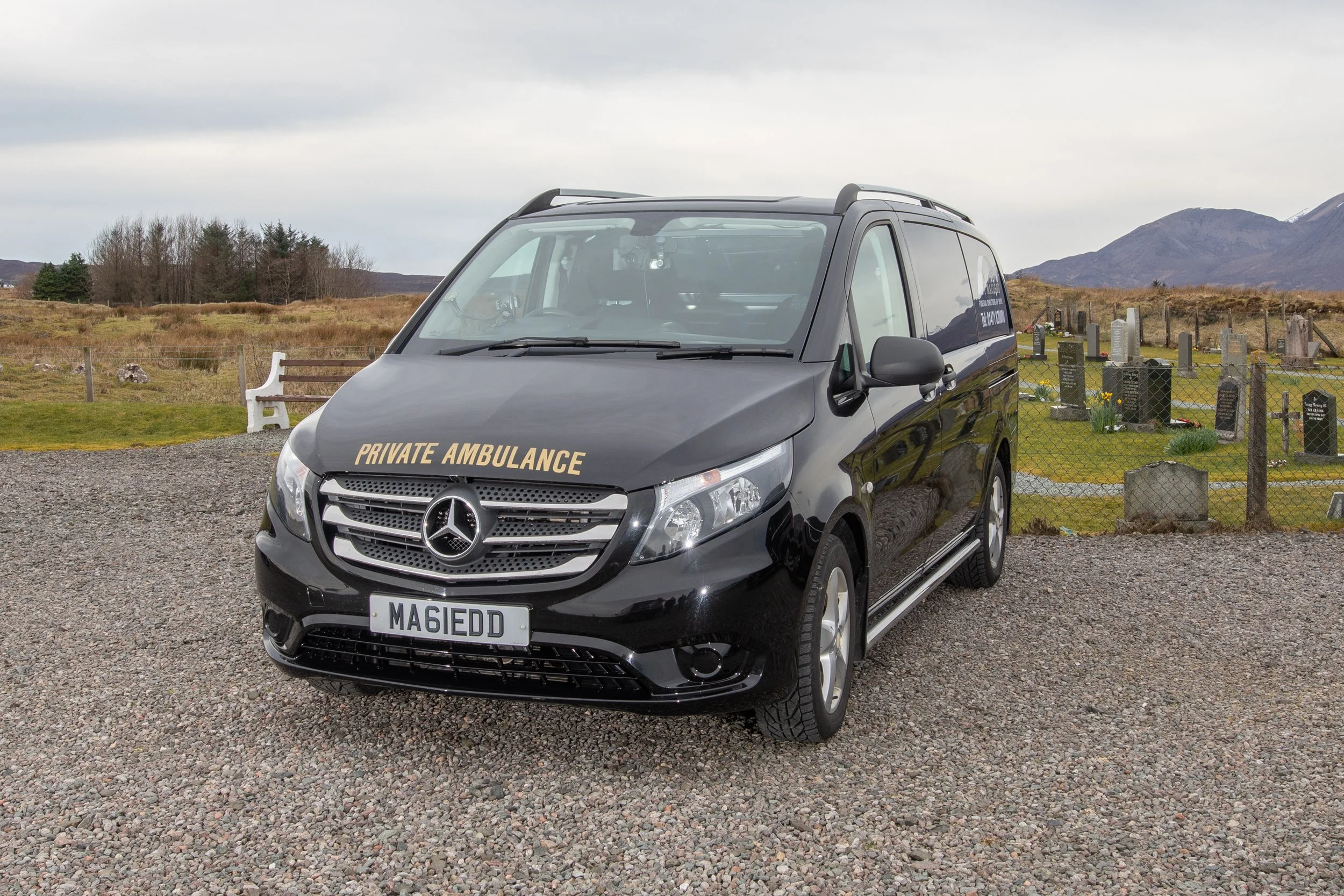 A black Mercedes-Benz private ambulance van parked on a gravel surface with a rural landscape, hills, and mountains in the background under an overcast sky.