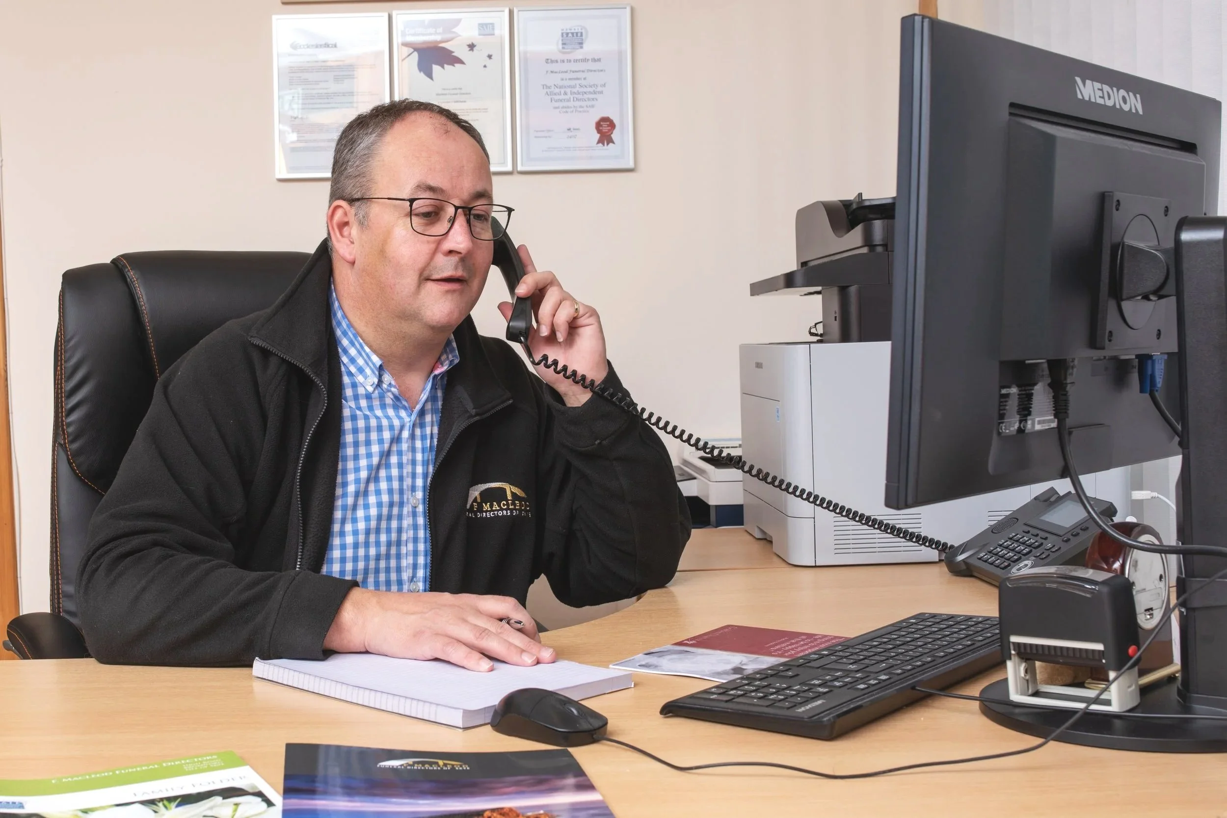 A man with glasses and a checked shirt sitting at a desk talking on a corded phone, with a computer monitor, keyboard, and various papers and brochures on the desk, in an office environment.
