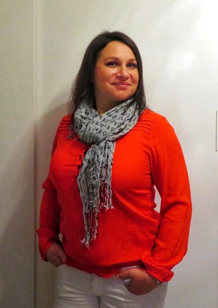 Eine Frau mit dunklem Haar, die ein rotes Oberteil und einen grauen Schal trägt, steht vor einer hellen Wand.