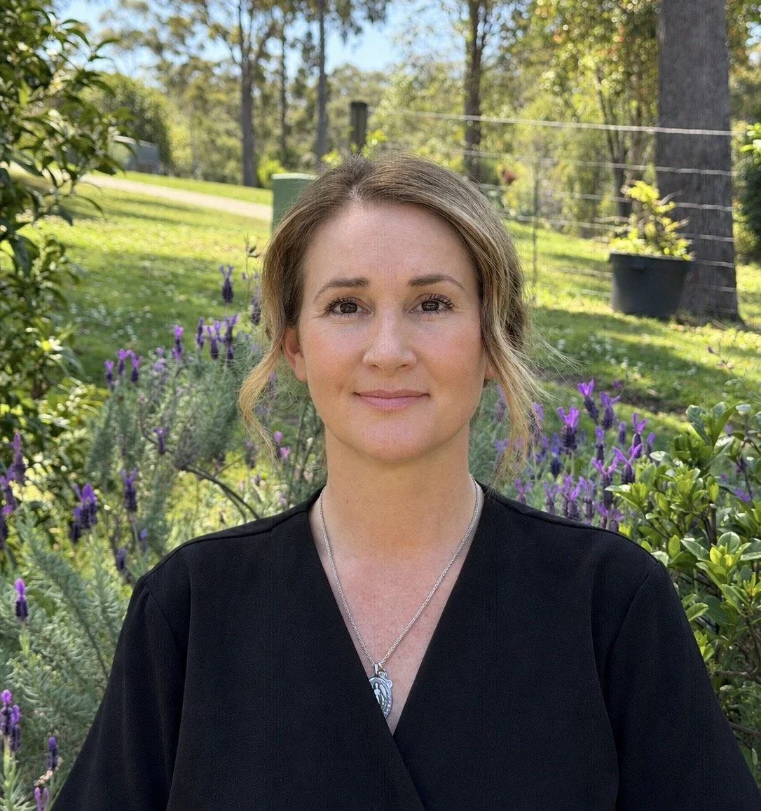 A woman with brown hair, wearing a black top and a silver necklace, standing outdoors in a garden with purple flowers and green trees in the background.