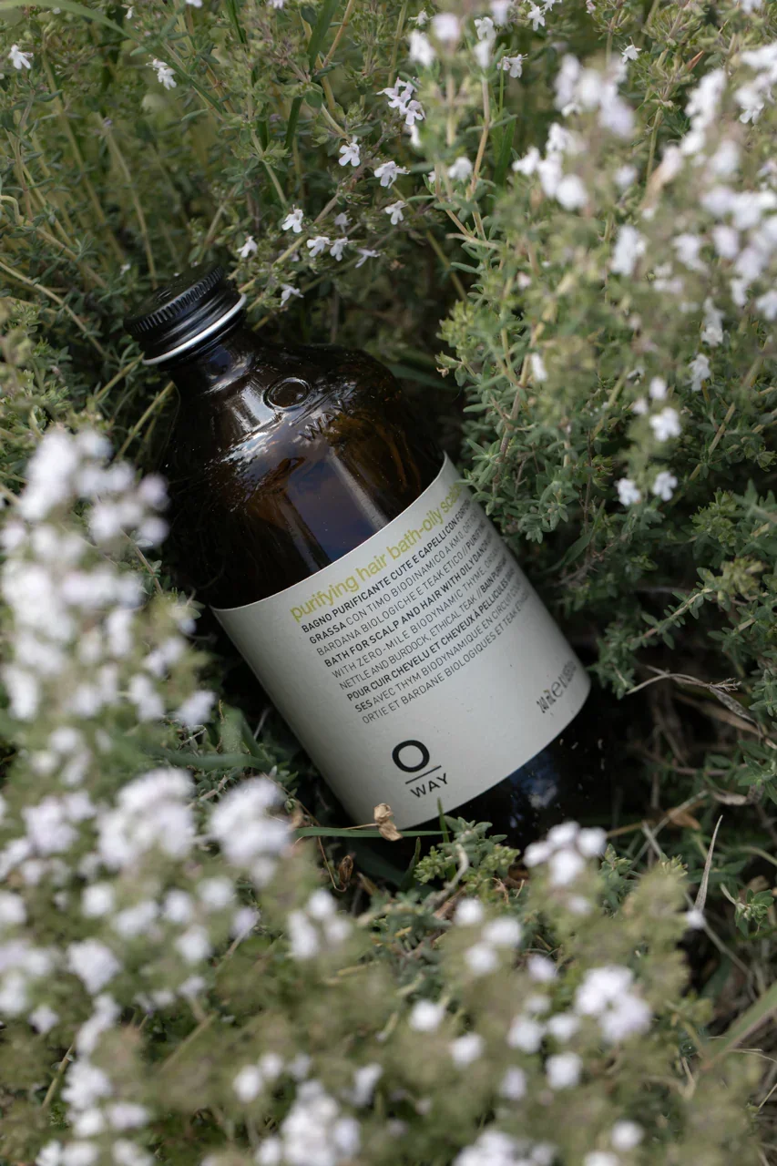 A brown glass bottle labeled 'Purifying Hair Bath' lying among green plants and small white flowers.