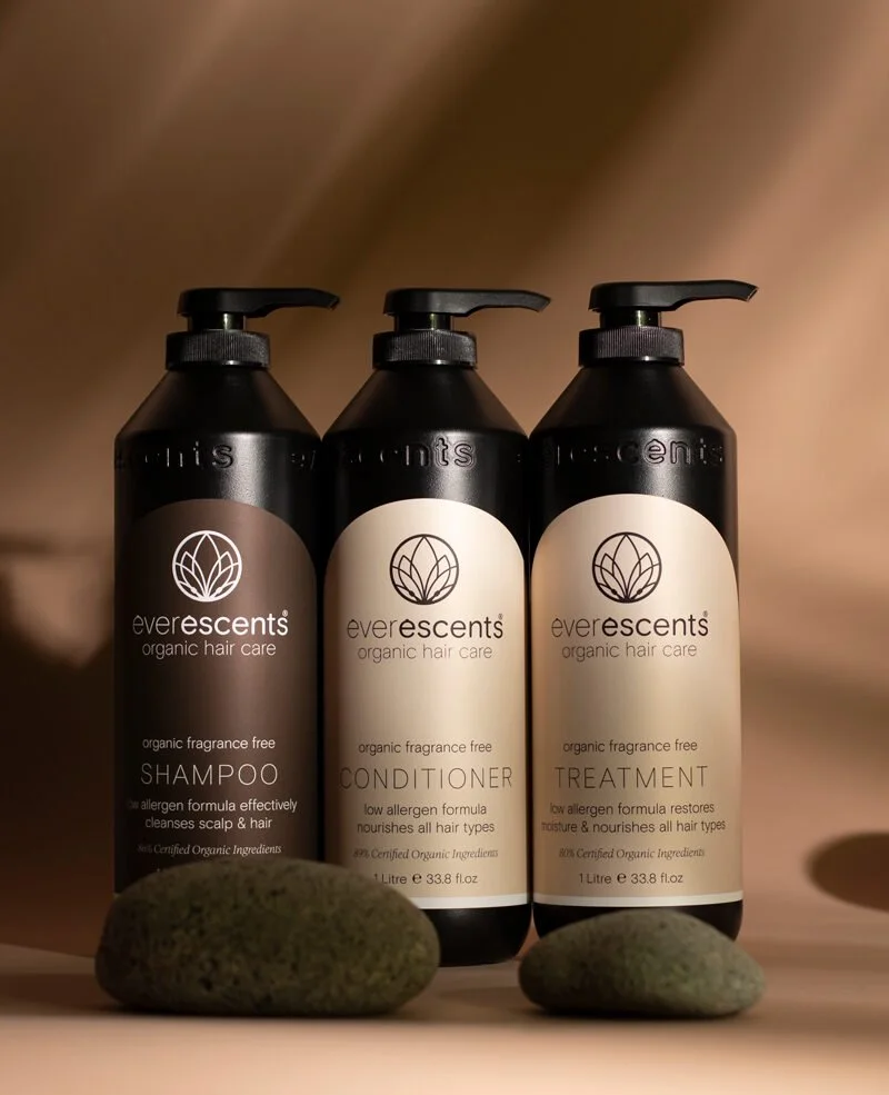 Three black bottles of Ever Escents organic hair care products, including shampoo, conditioner, and treatment, arranged in a row on a surface with two rocks in front of them.