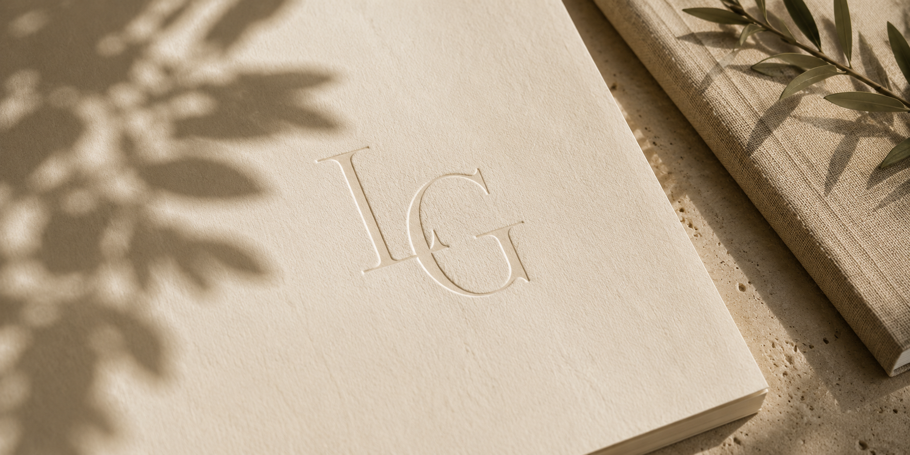 Cream-colored textured card with embossed initials I & G, shadowed by a leafy branch, placed on a beige surface alongside a textured beige fabric and a closed book.