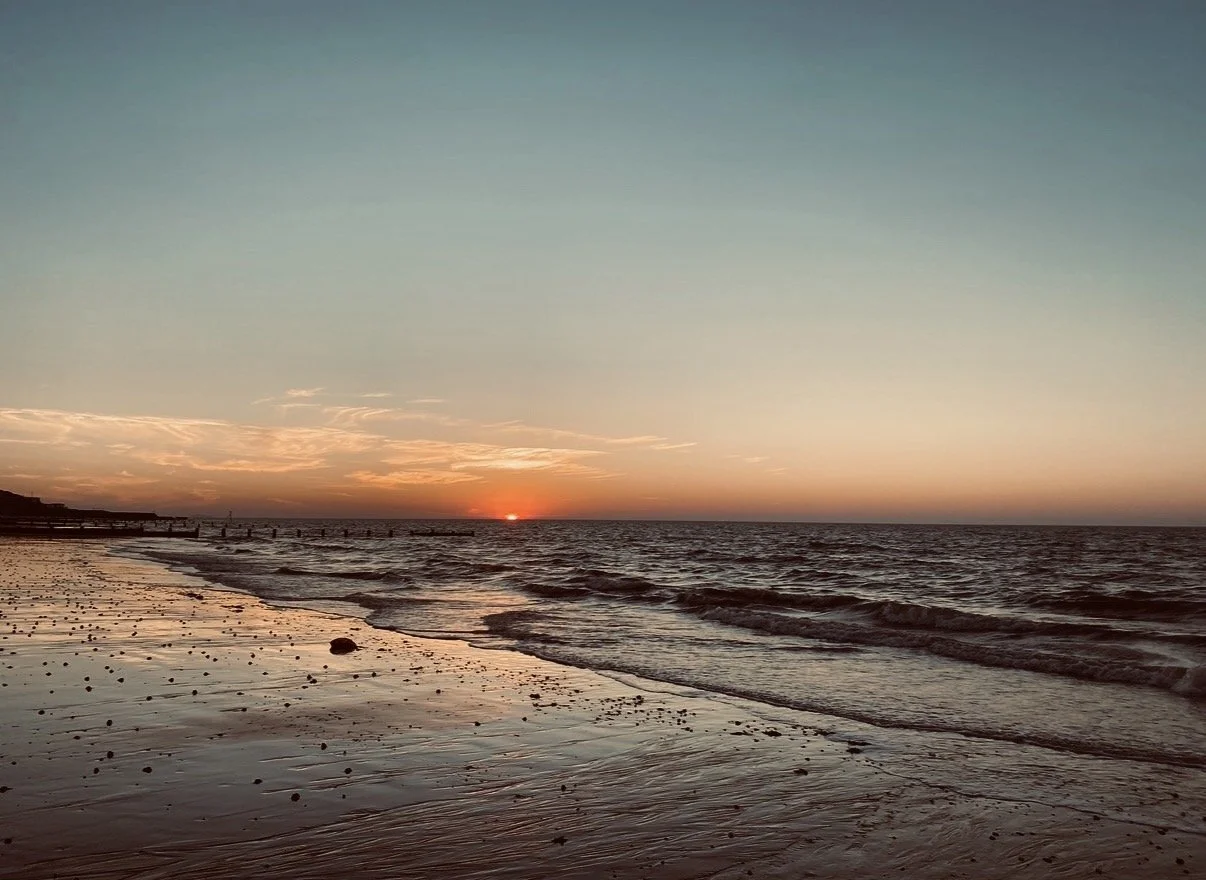 Sunset over the ocean with a sandy beach in the foreground, gentle waves, and a pier extending into the water on the left.