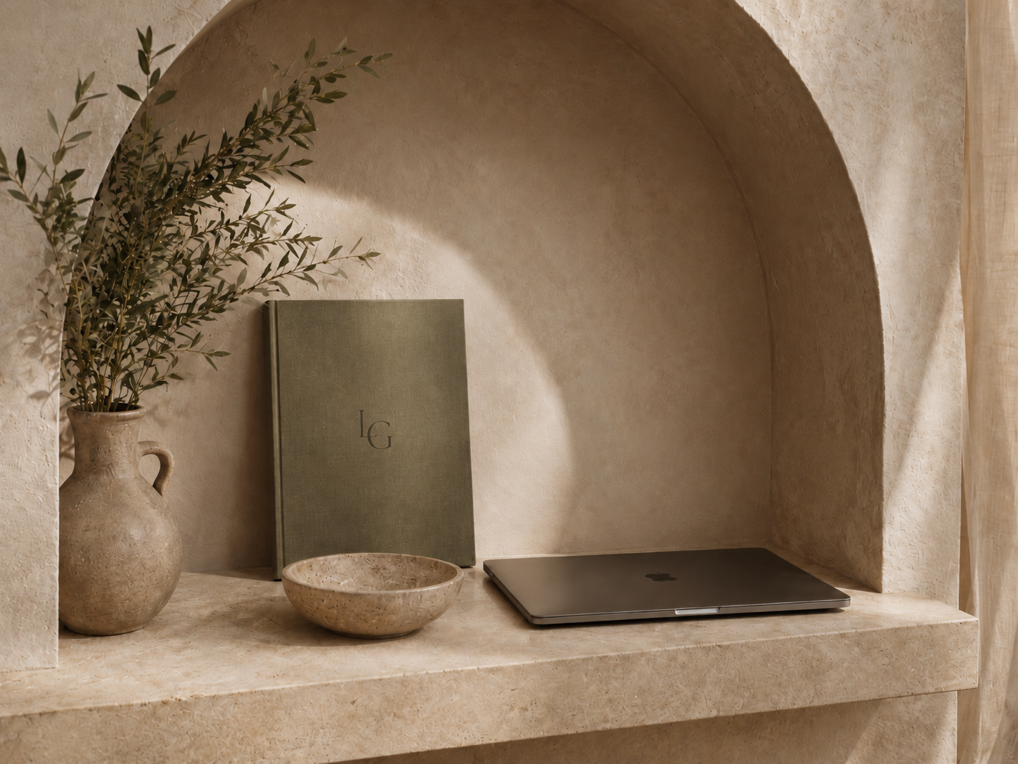 A beige marble shelf built into a wall niche, holding a beige vase with olive branches, a green book with initials 'L G', a small beige bowl, and a closed silver laptop.