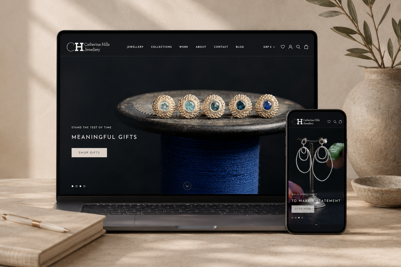 Laptop and smartphone displaying the website of Catherine Hills Jewellery, featuring jewelry products like rings and earrings, placed on a wooden desk with a notebook and pen, and a vase with a plant in the background.