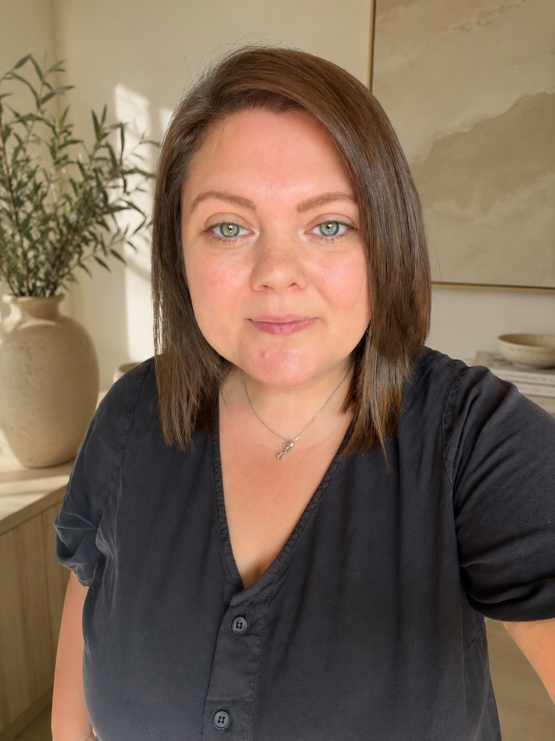 A woman with shoulder-length brown hair and light blue eyes taking a selfie in a modern, well-lit room. She is wearing a black shirt and a silver necklace with a treble clef pendant. In the background, there is a large beige vase with green foliage, a piece of abstract artwork, and a table with bowls.