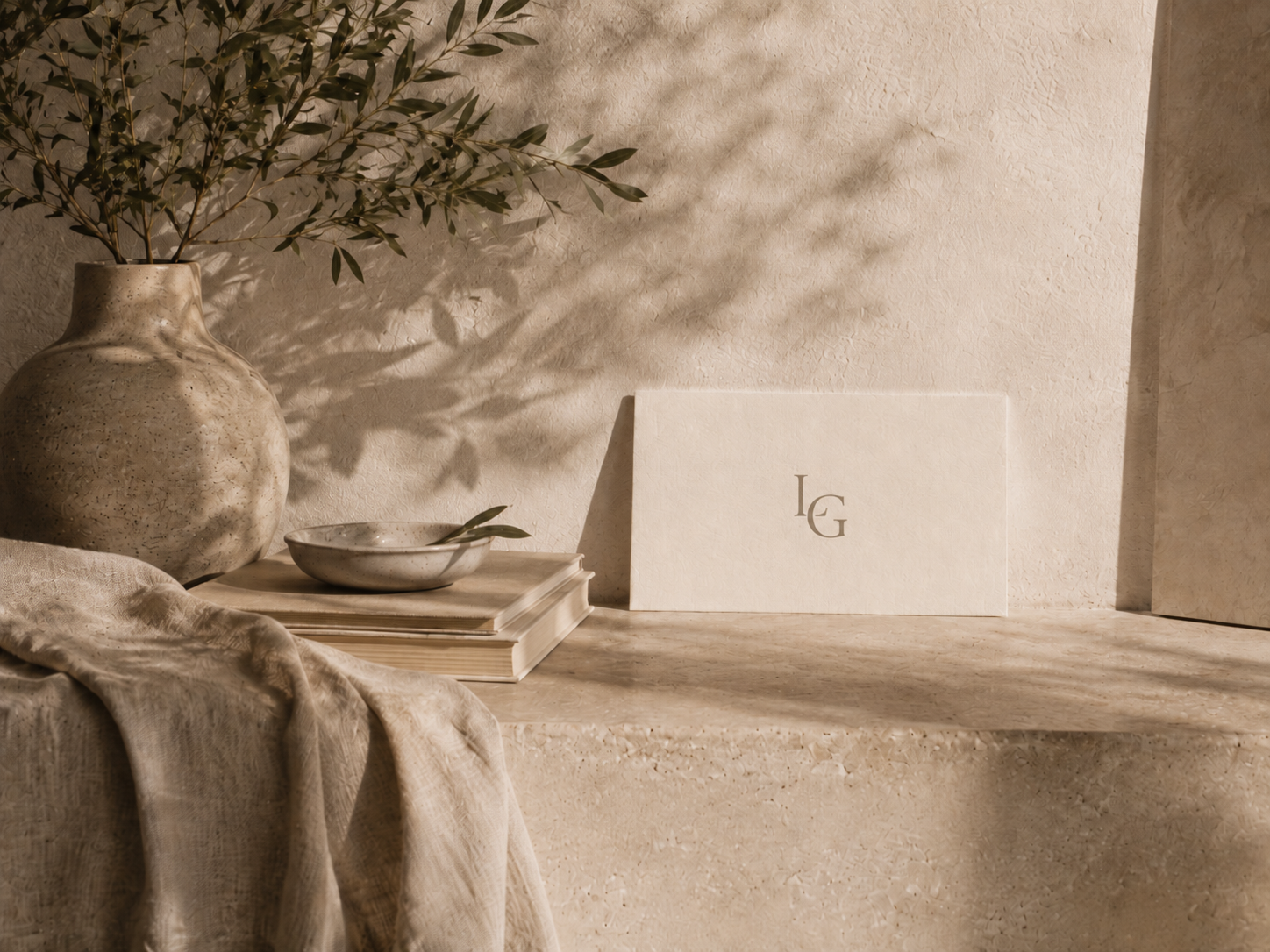A beige textured vase with green leafy branches, a small bowl with a single leaf, a stack of books, and a framed print with initials 'L G' on a neutral stone surface with a light-colored cloth in a minimalistic interior setting.