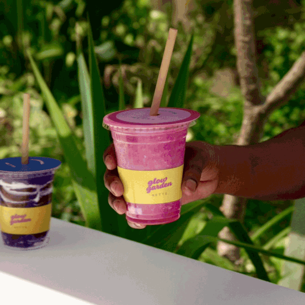 Close-up of a hand holding a pink smoothie with a straw, on a table with a similar drink in a transparent cup with a purple lid, outdoors with green plants in the background.