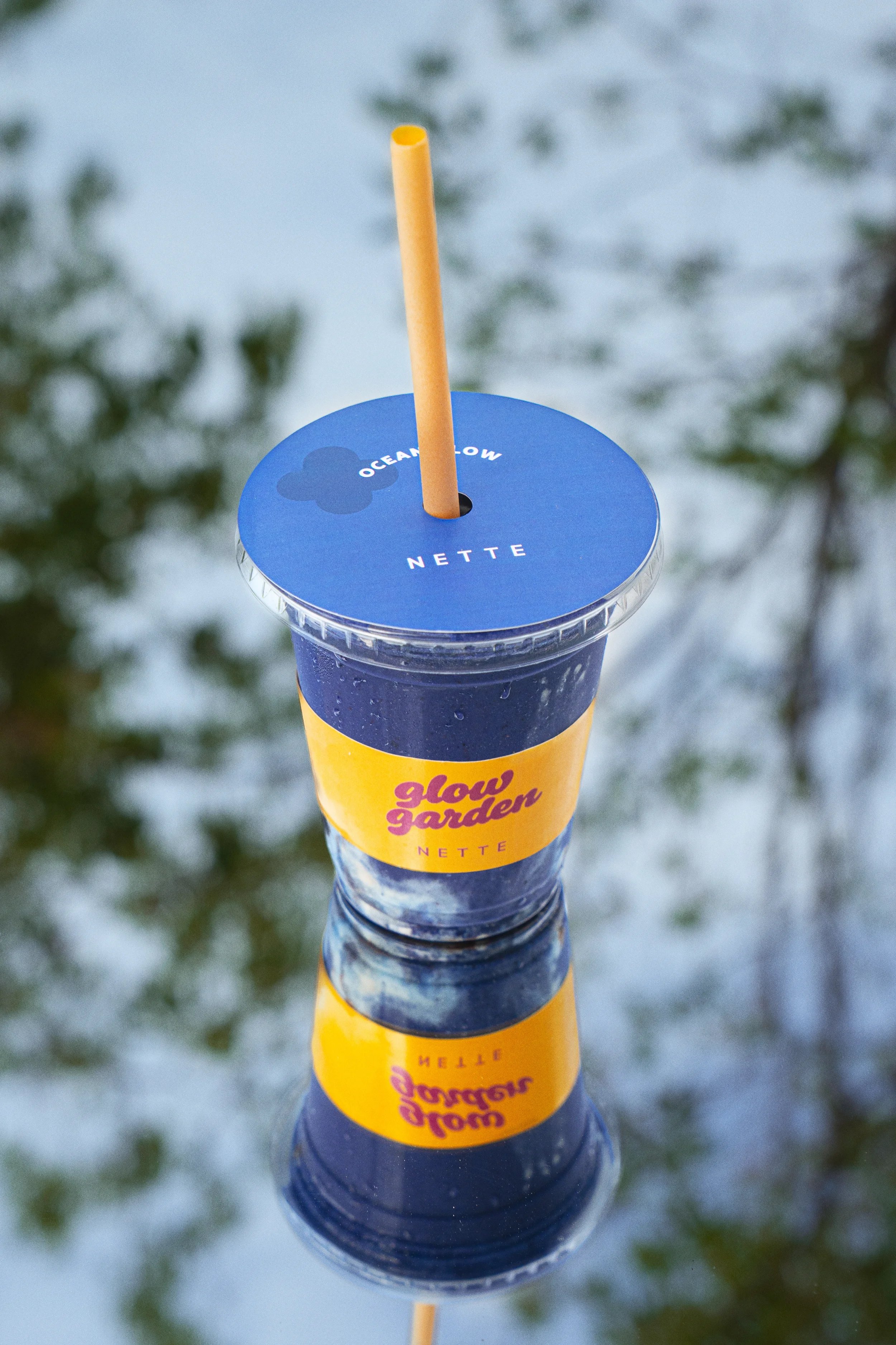 Close-up of a blue plastic cup with a yellow straw, featuring a yellow band with pink and purple text that says 'Glow Garden,' on a blurry background of snow and trees.