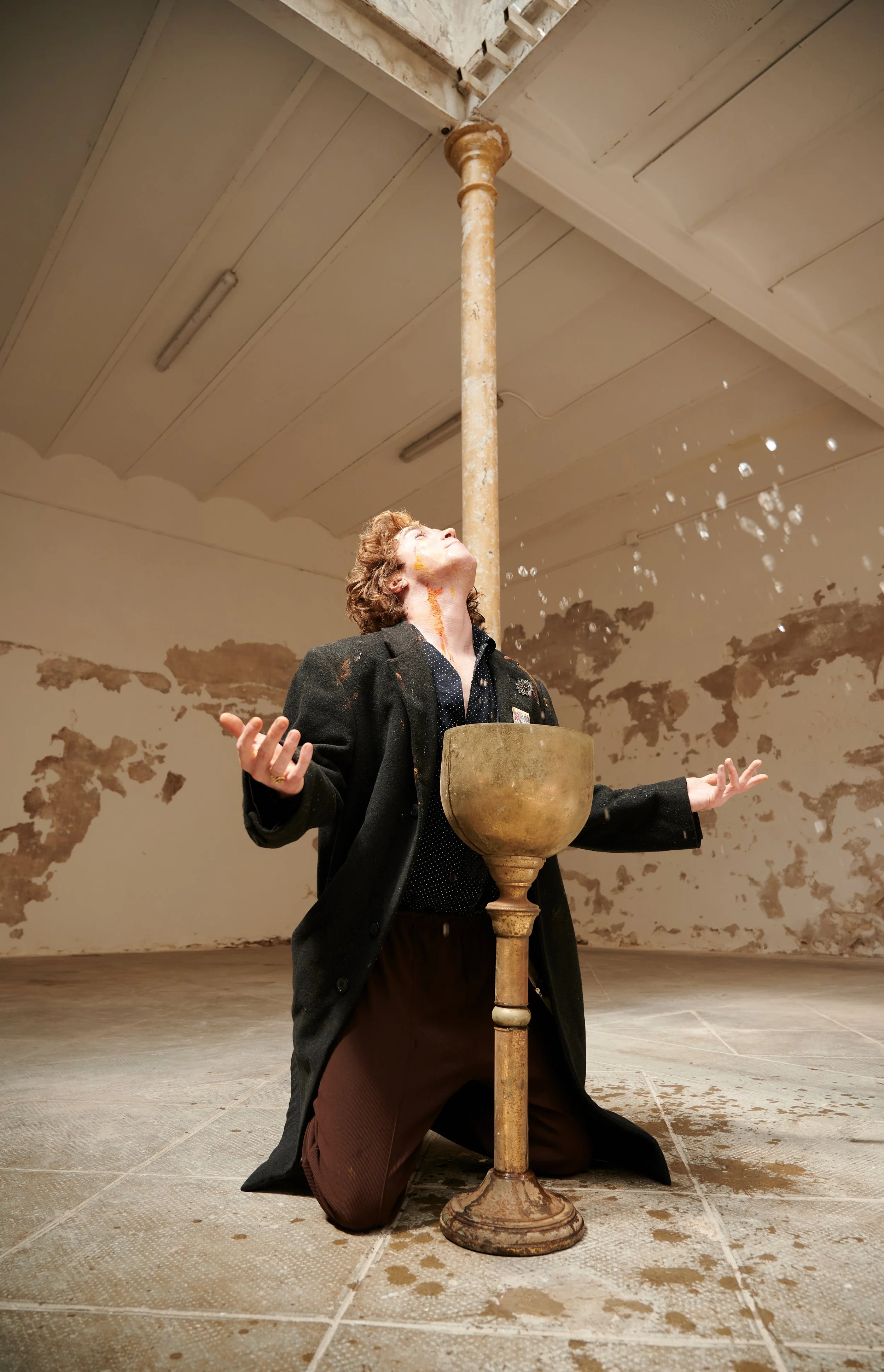 A person is kneeling on the floor with arms raised, head tilted back, and eyes closed, in front of a large, old, tarnished brass candlestick holder with a bowl-shaped top, in an empty room with peeling paint and a rustic atmosphere.