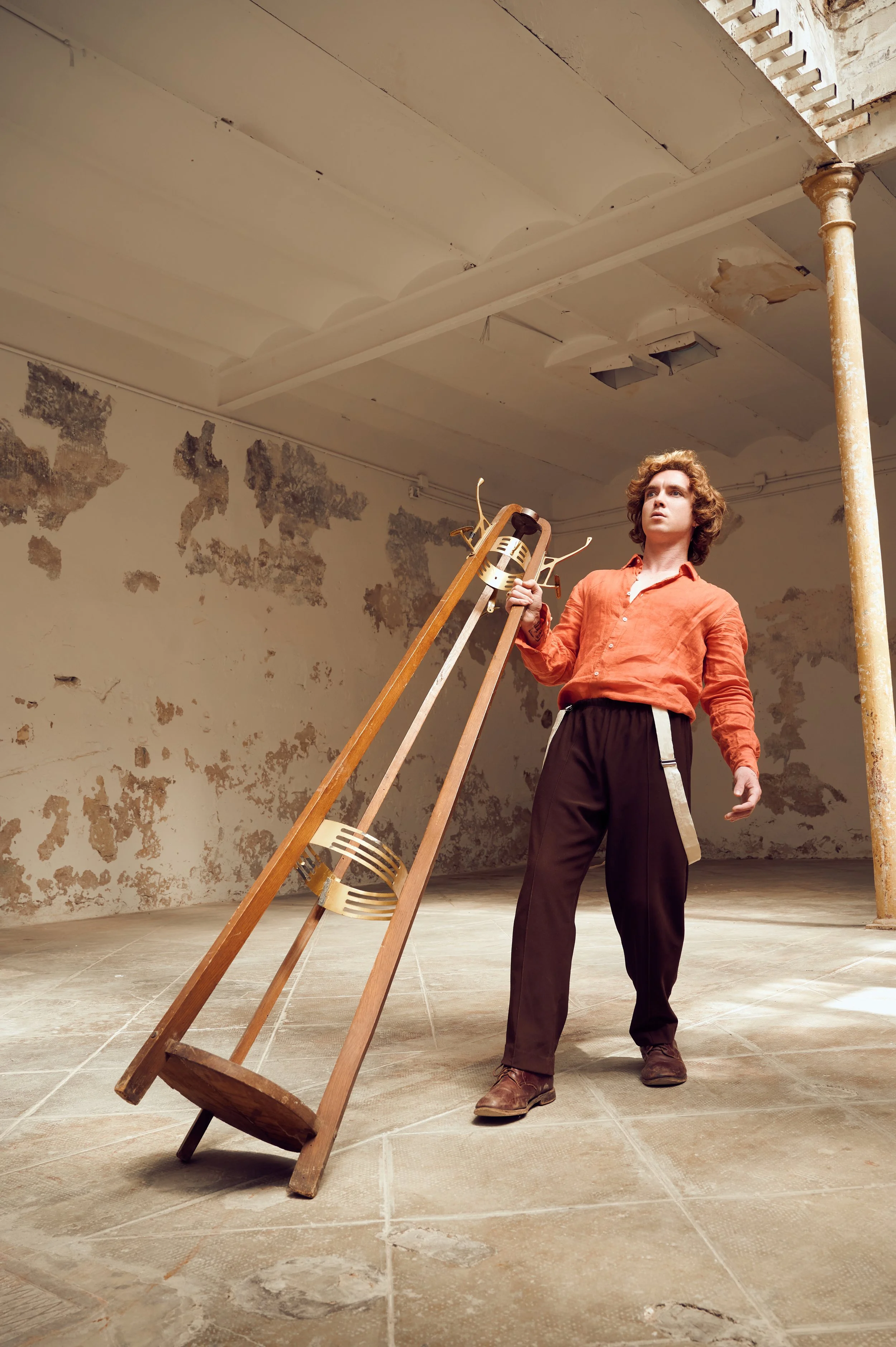 A young man with curly hair, wearing an orange shirt and dark pants, stands in a room with peeling walls and a cracked ceiling. He holds a wooden chair and stands near an angled wooden ladder.