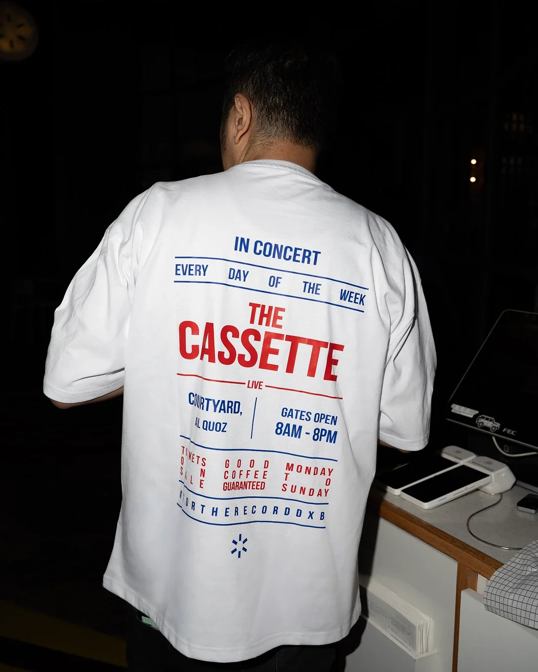 Back of a person wearing a white shirt with colorful text about a live music event called The Cassette, including details about schedule and tickets.