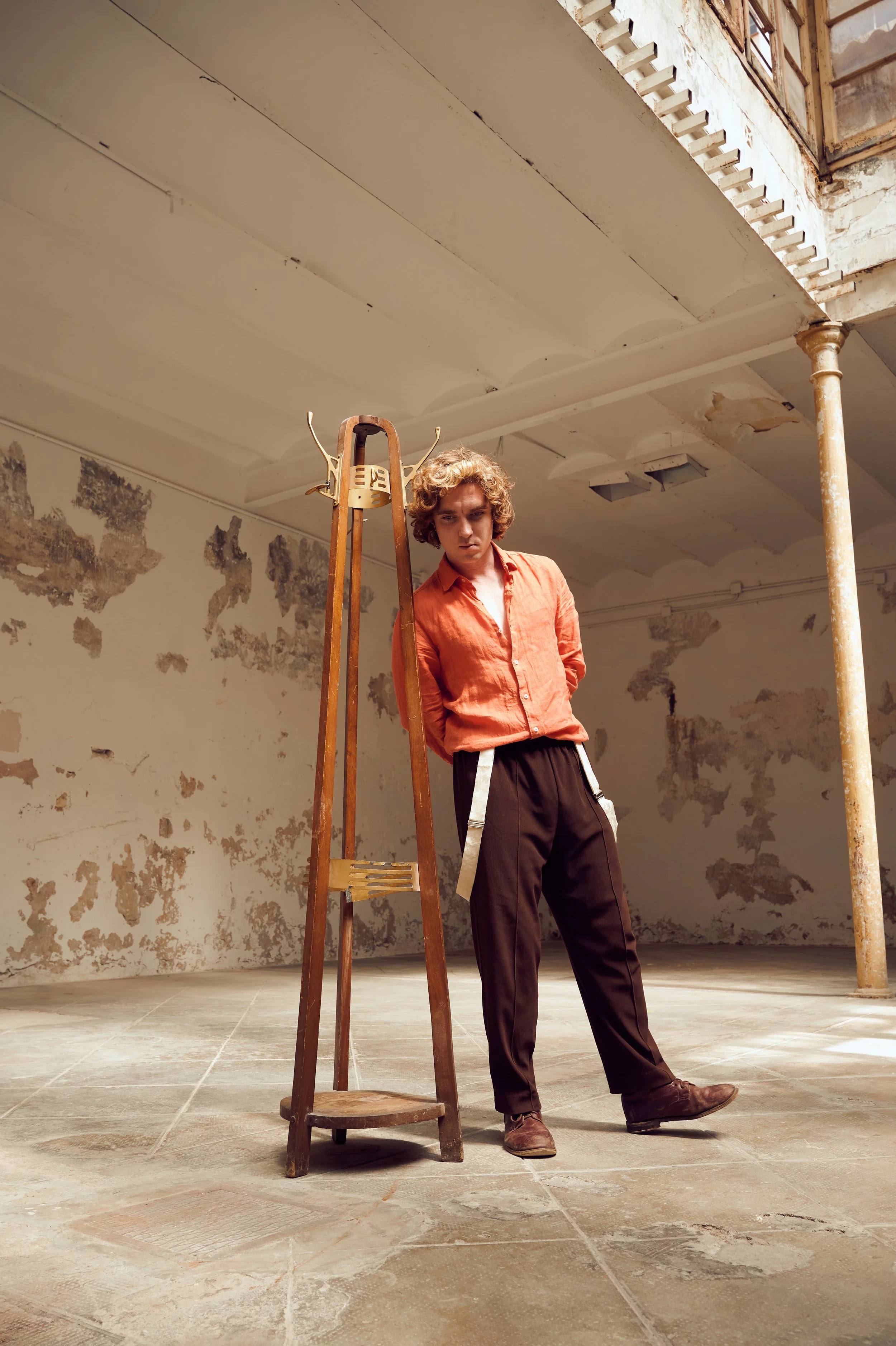 A young man with curly hair wearing an orange shirt and dark pants stands in an abandoned industrial space with peeling paint on the walls. He leans to the side on a wooden ladder, which is positioned in front of him. The space has high ceilings with