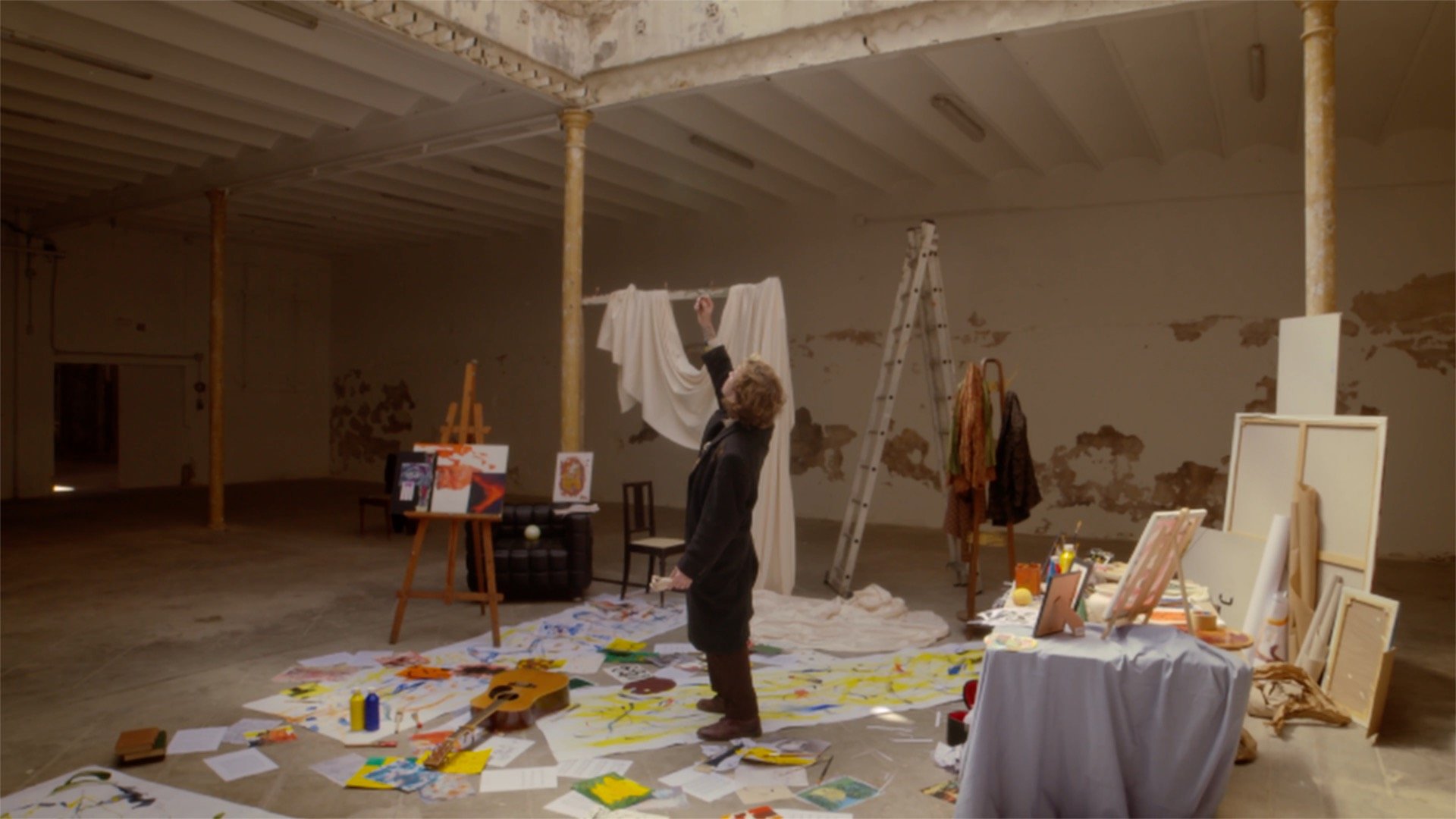 An art studio with a woman painting on a large canvas on the floor, surrounded by paintings, art supplies, and canvas frames. The room has scaffolding and a ladder, with peeling wall paint.