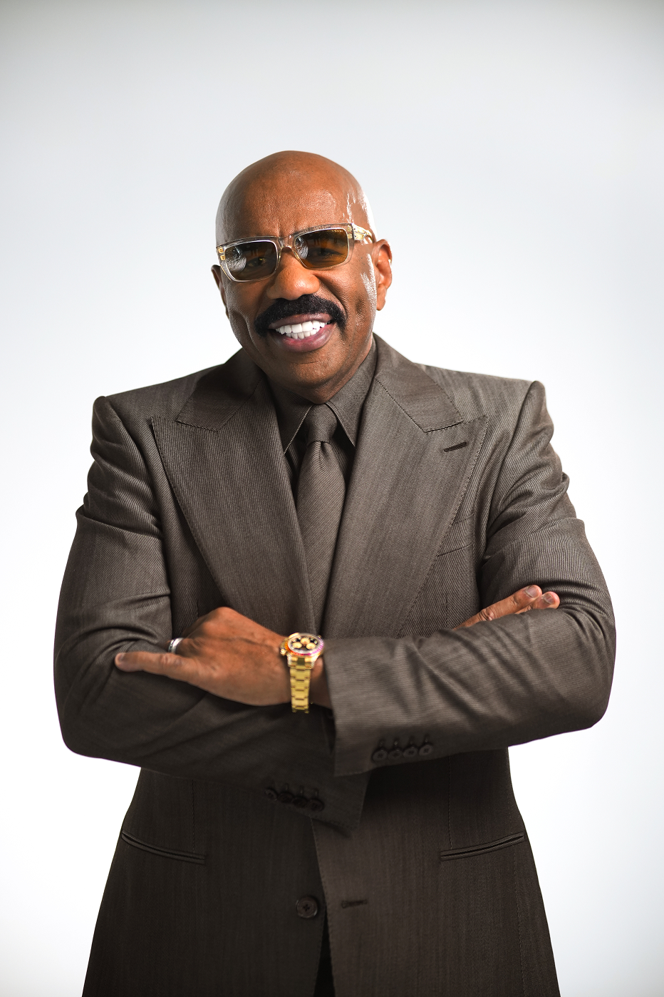 A smiling man with sunglasses and a mustache wearing a brown suit, crossing his arms, standing against a plain white background.