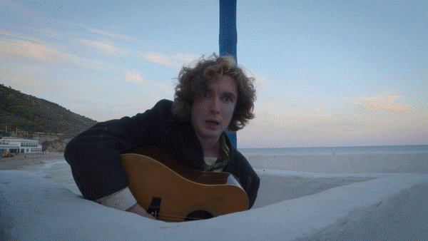 A woman with curly hair playing an acoustic guitar outdoors near the water, with a beach and hills in the background.