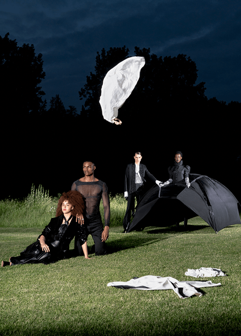 Four models outdoors at night, one sitting and three standing, with a large black umbrella and clothing on the grass, under a dark sky with a white cloth in the shape of a wing hanging above.