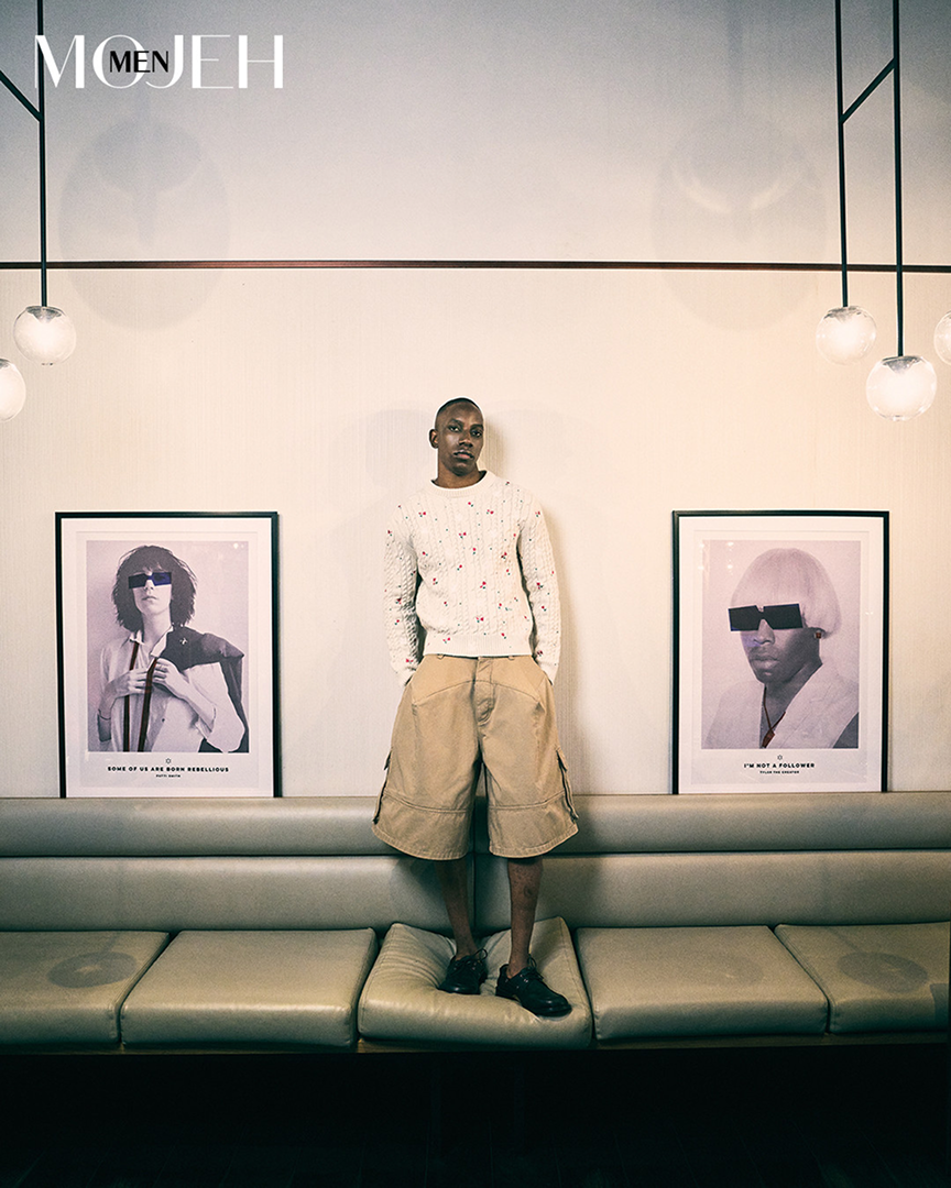 A person standing on a beige cushioned bench in a modern room with framed black-and-white portraits on the wall behind them. The person has a shaved head and is wearing a white sweater with colorful spots, oversized beige shorts, and black shoes.
