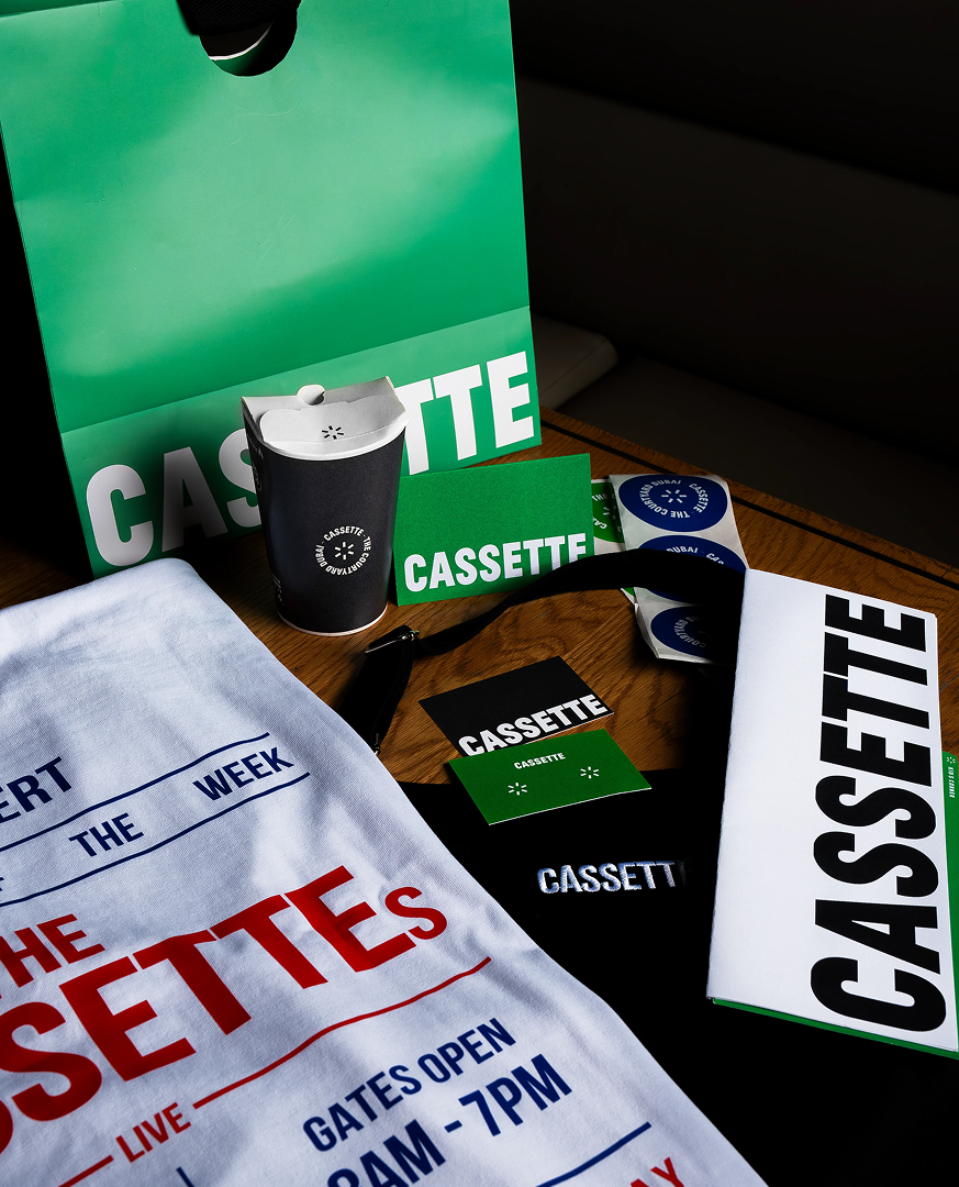 Cassette-themed promotional items including stickers, a wristband, a small paper cup with a lid, and signs, arranged on a table with a green and white Cassette bag and a white cloth with event information for a concert or event.